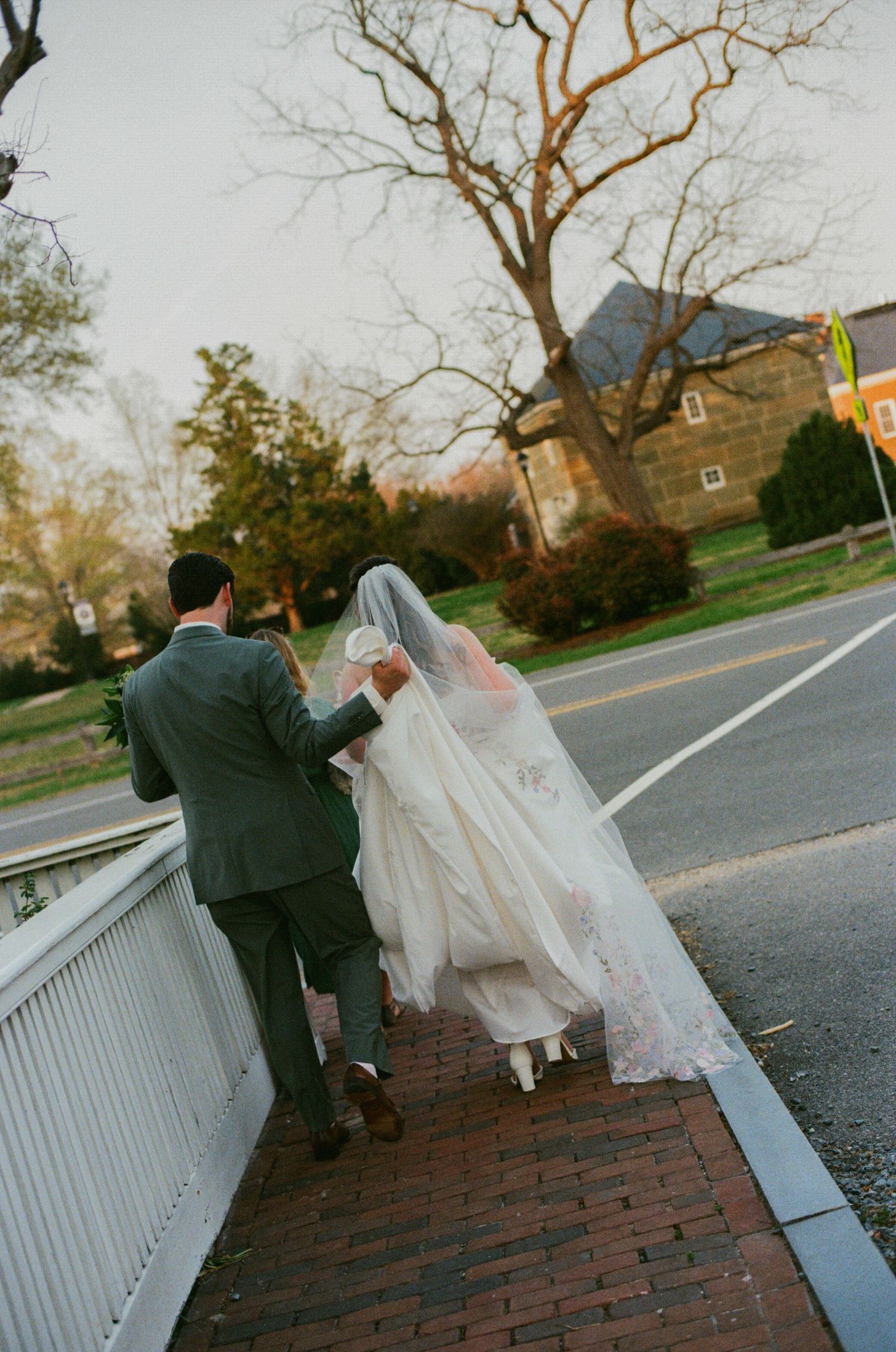 Bride and groom walking hand in hand on a brick sidewalk near a road, with trees and a stone building in the background.