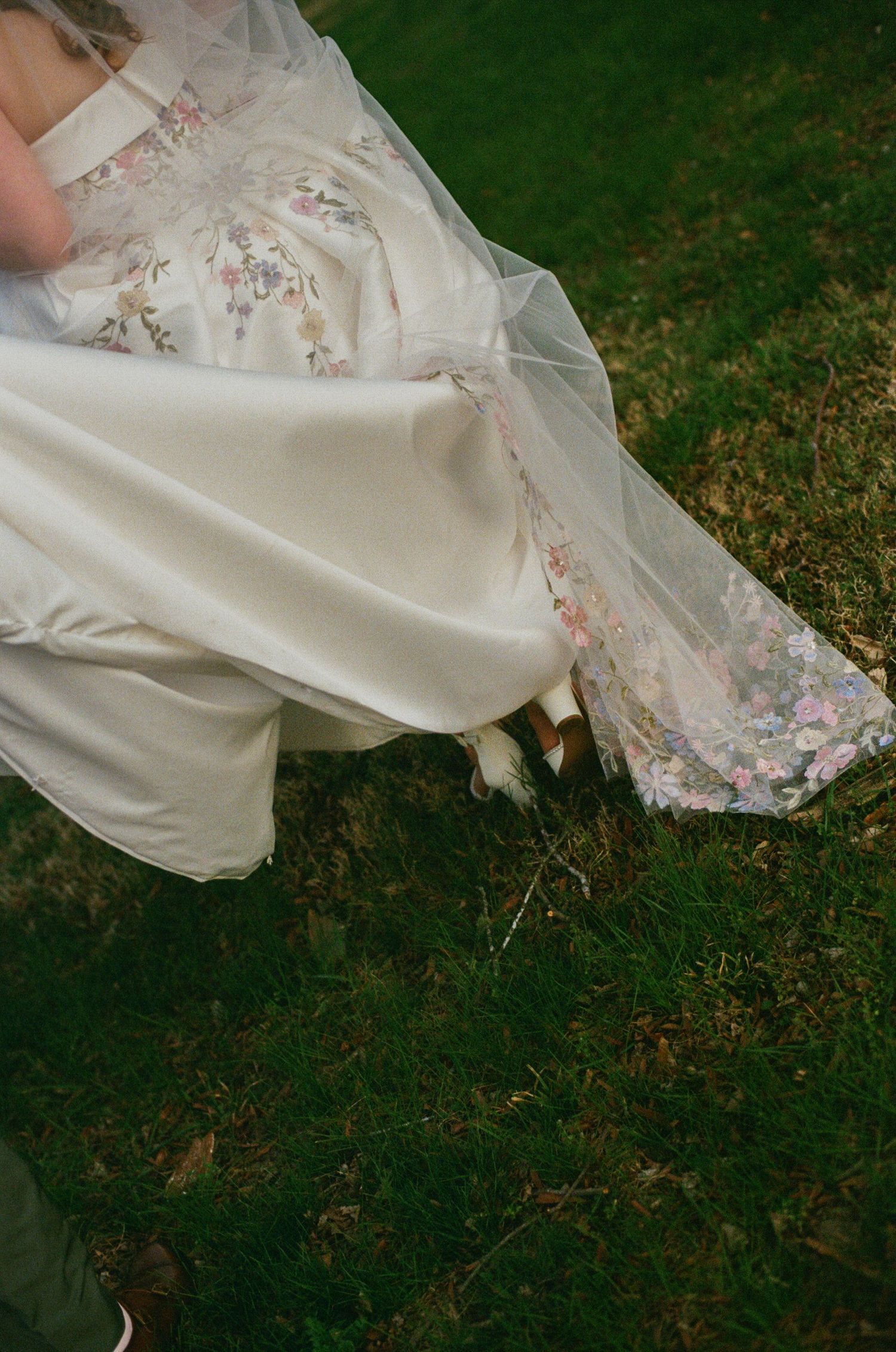 Bride in floral wedding gown with veil standing on green grass, showcasing elegant dress details.