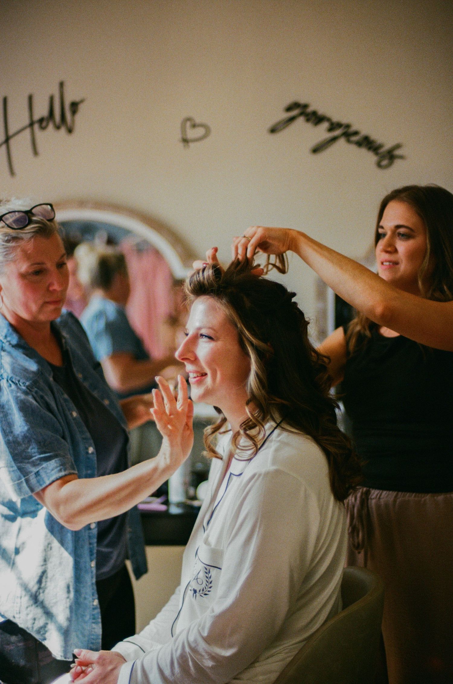 Bride smiling while two women style her hair in a bright salon with Hello and Gorgeous wall decor.
