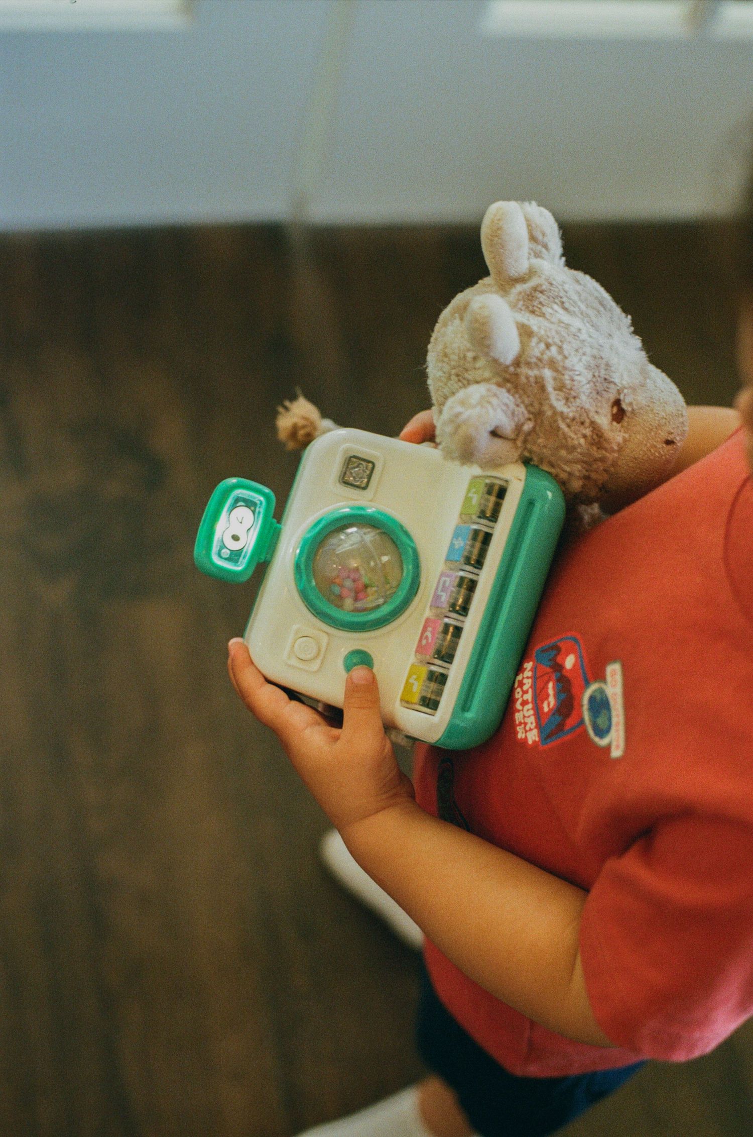 A young child holds a Fisher-Price toy camera and a stuffed teddy bear while wearing a red shirt.