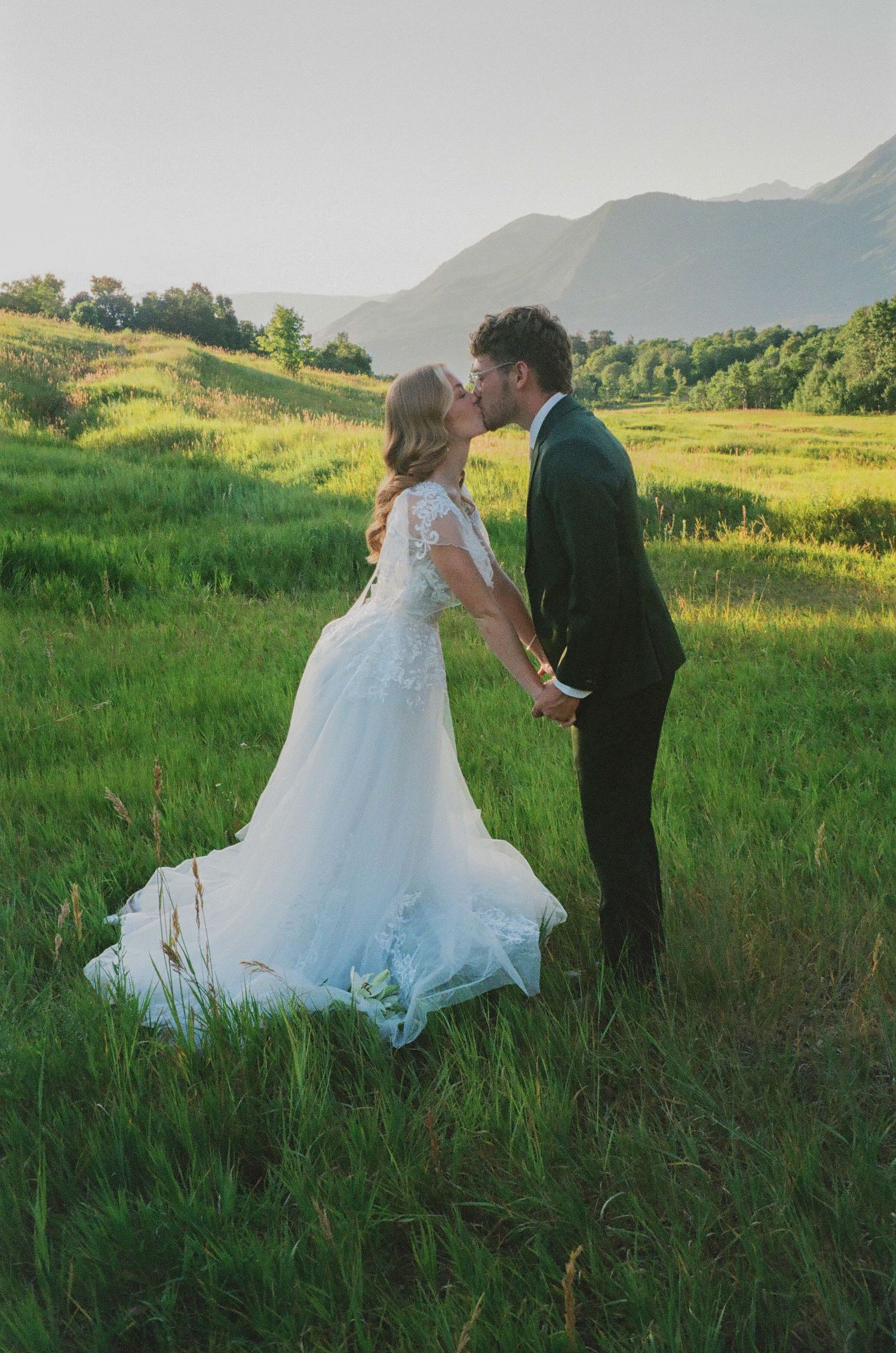 Bride and groom kissing in a lush green meadow with mountain backdrop during golden hour wedding photography.