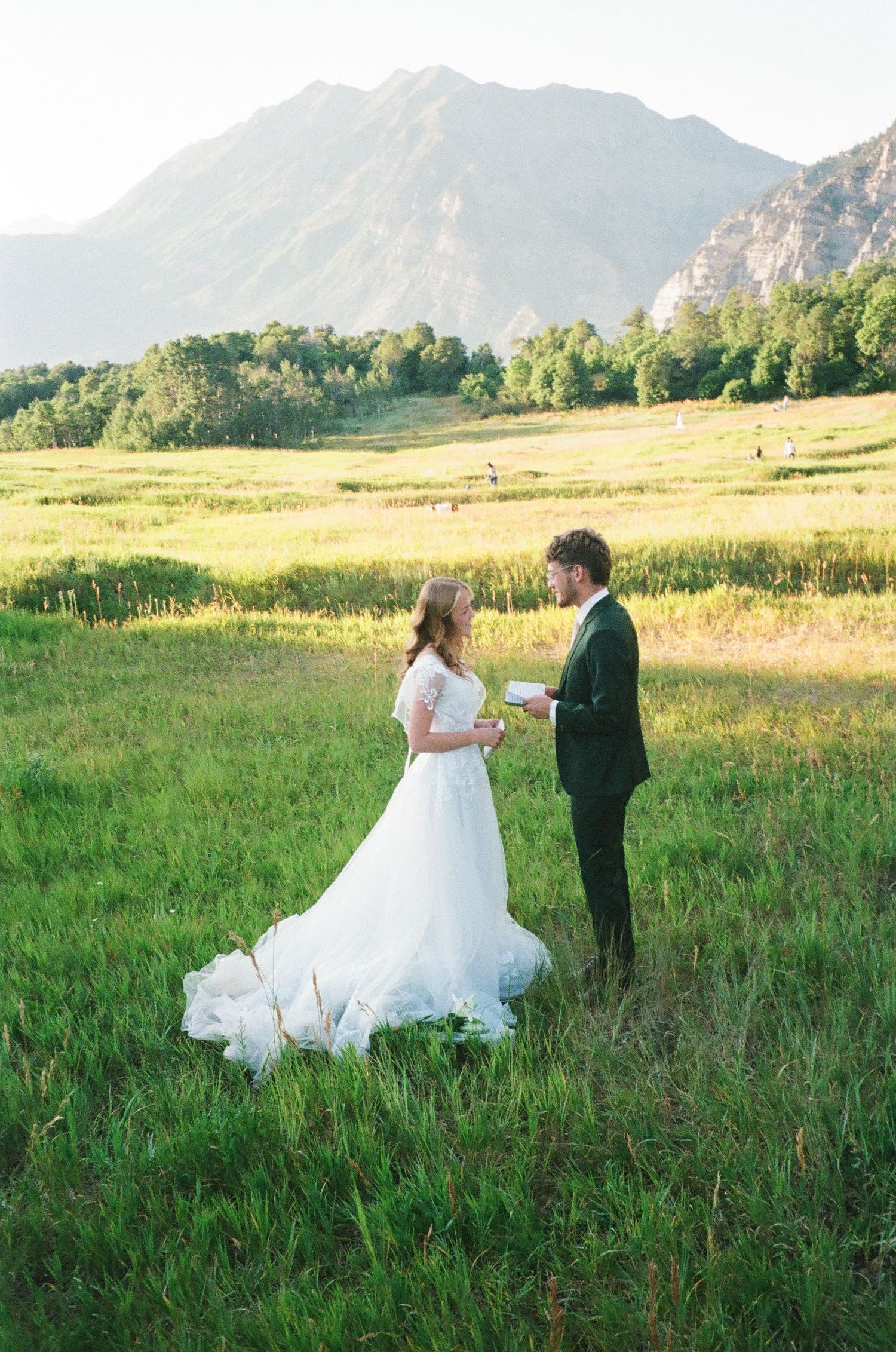 Bride and groom exchange vows in a lush green mountain meadow, bride wearing elegant white gown with long train.