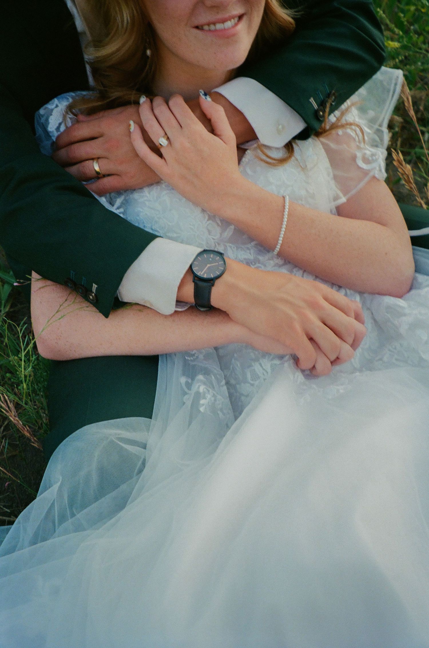 Bride in white lace dress and groom in green suit embracing outdoors, showing wedding rings and a black wristwatch.