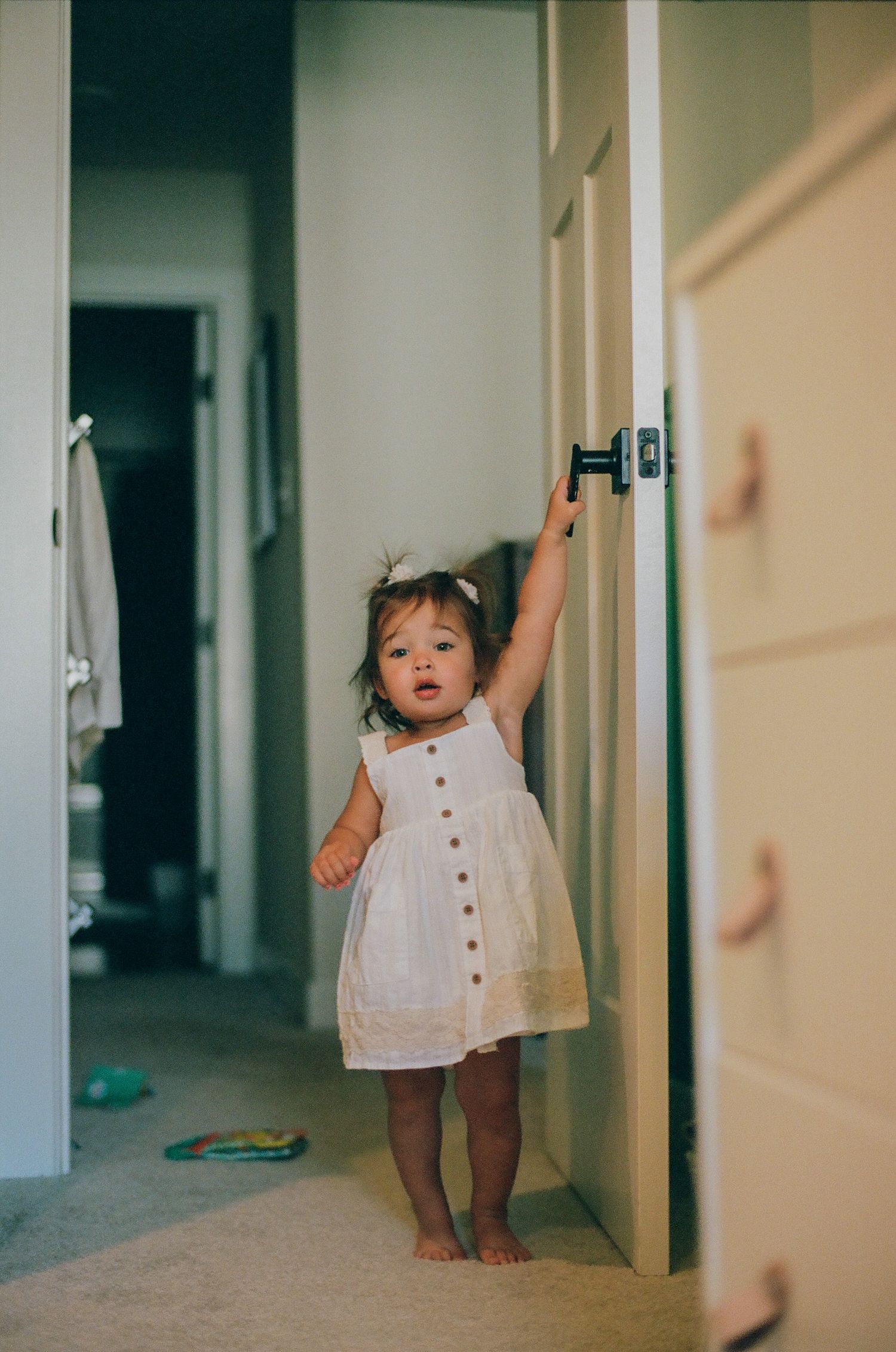 Toddler girl in white button dress reaching up to open a door handle in a home hallway.
