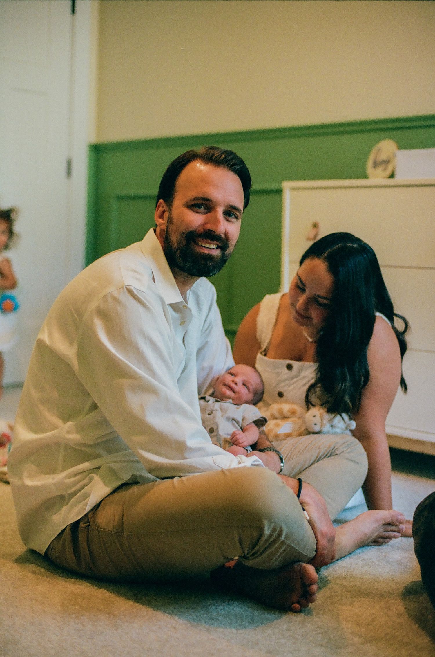 A couple sits on the floor smiling, cradling their newborn baby in a cozy green nursery room.