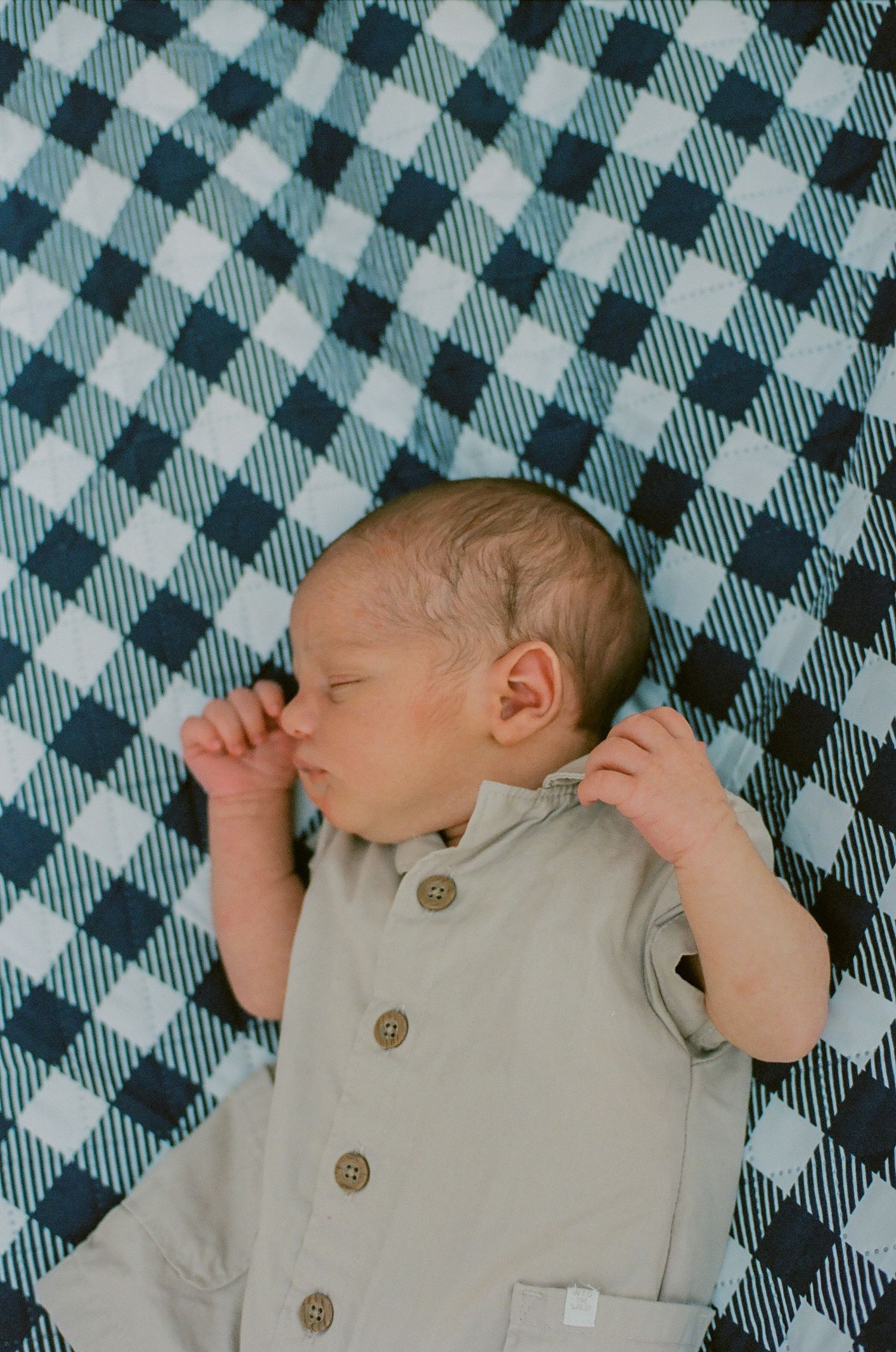 Sleeping newborn baby in sage green button romper lying on blue gingham blanket.