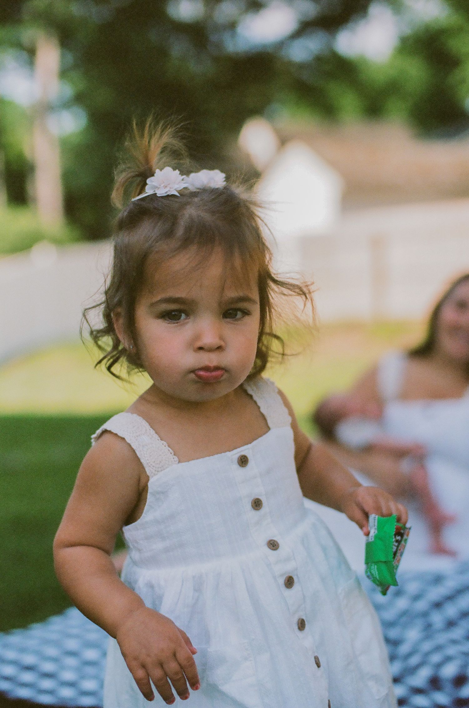 Adorable toddler girl in a white button dress with a floral hair clip holds a green toy outdoors.