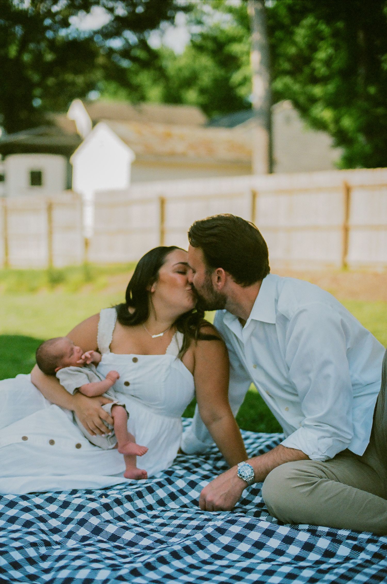 A couple shares a tender kiss while sitting on a gingham blanket outdoors, cradling their newborn baby.