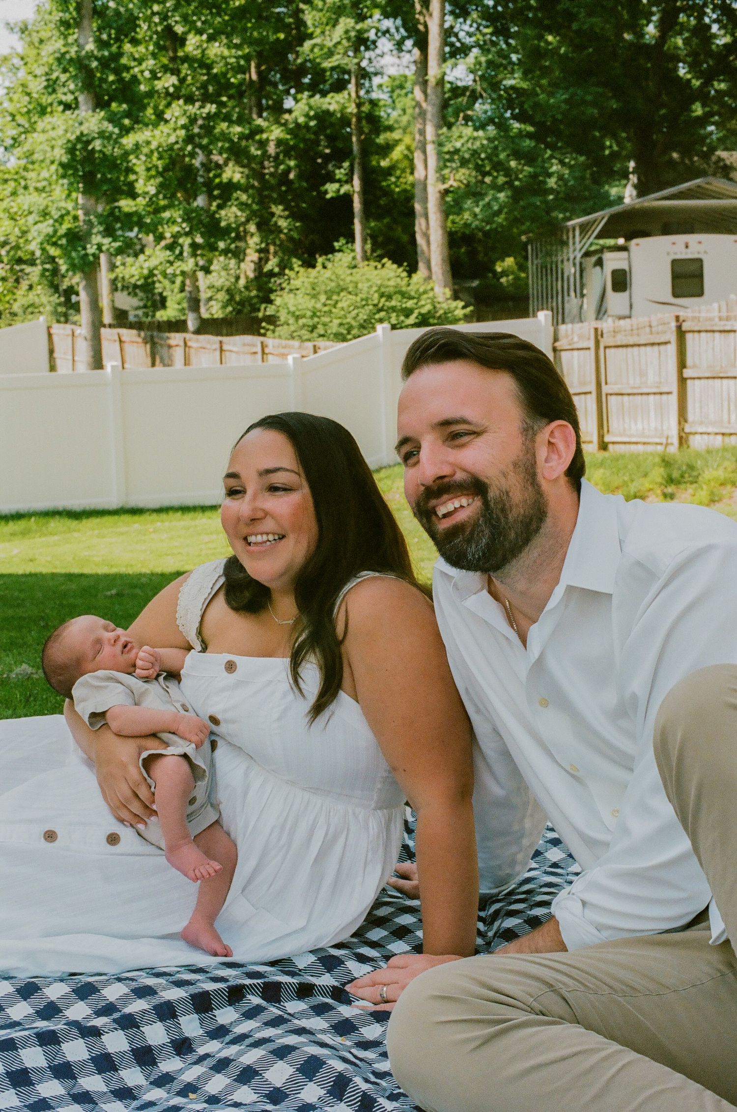 Happy couple sitting on a picnic blanket outdoors, smiling while holding their newborn baby in a backyard.