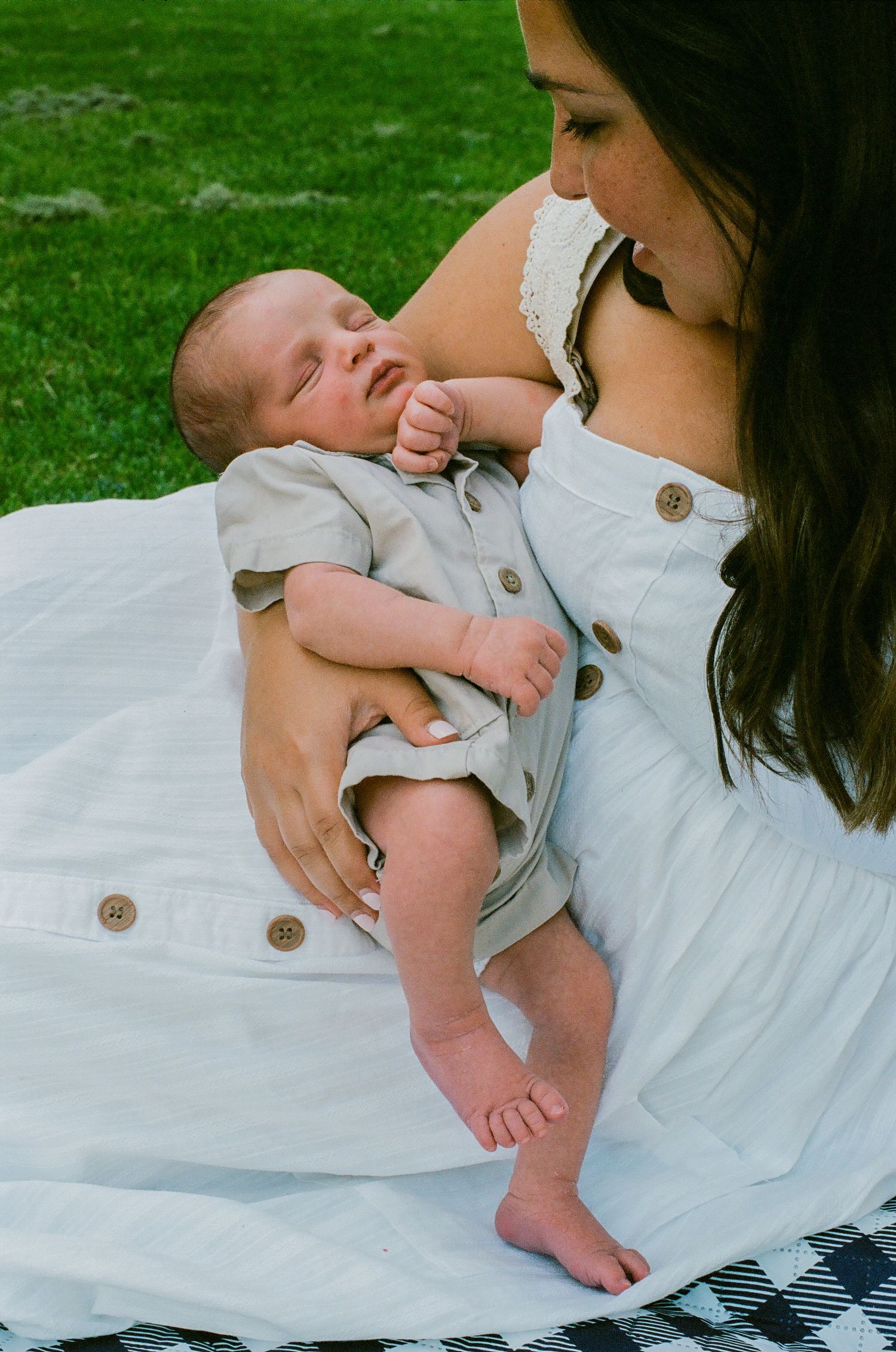 A mother in a white dress holds her sleeping newborn baby outdoors on a blanket with green grass behind.