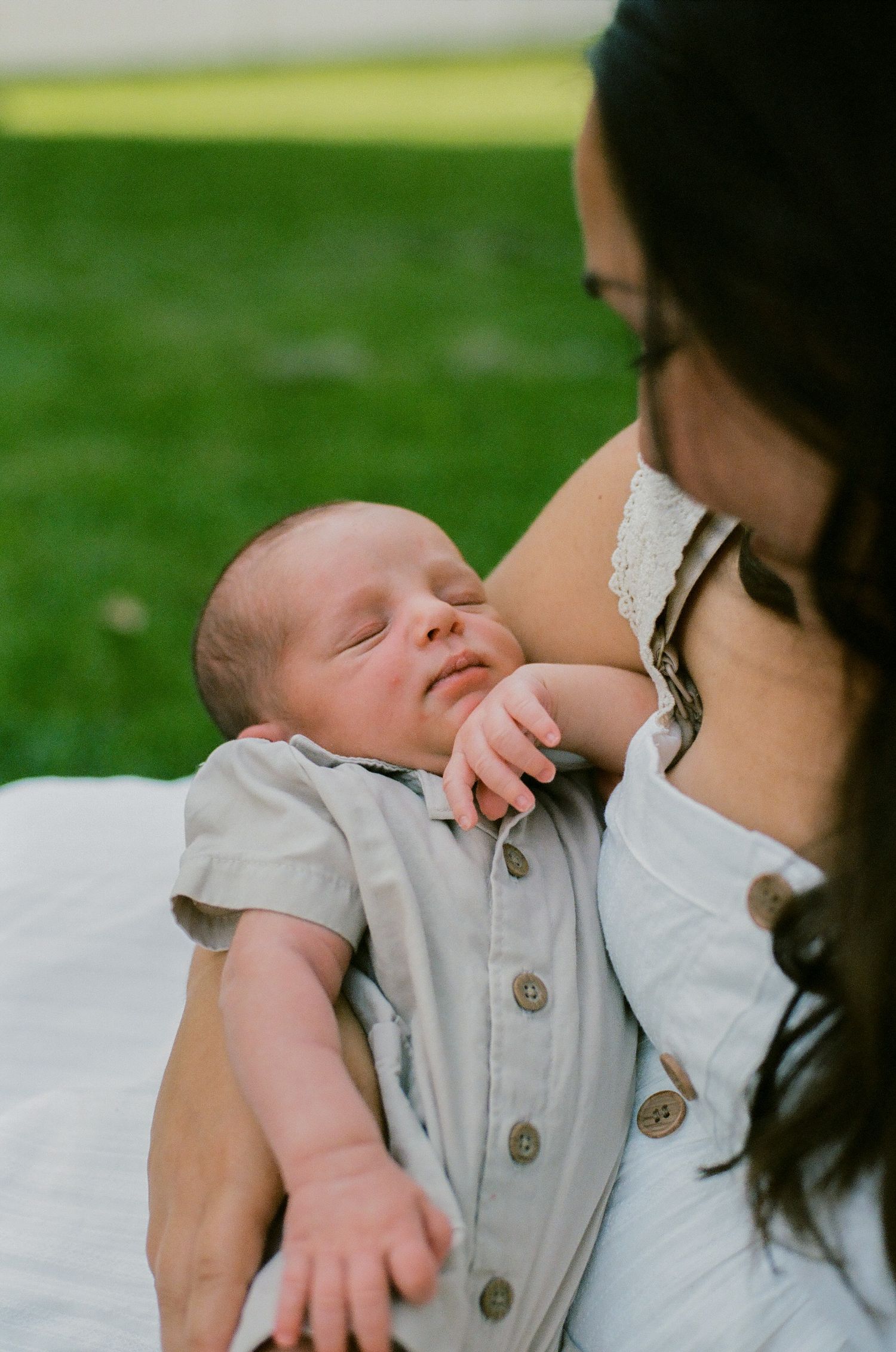 Close-up of a newborn baby in a gray romper resting peacefully in mothers arms outdoors.