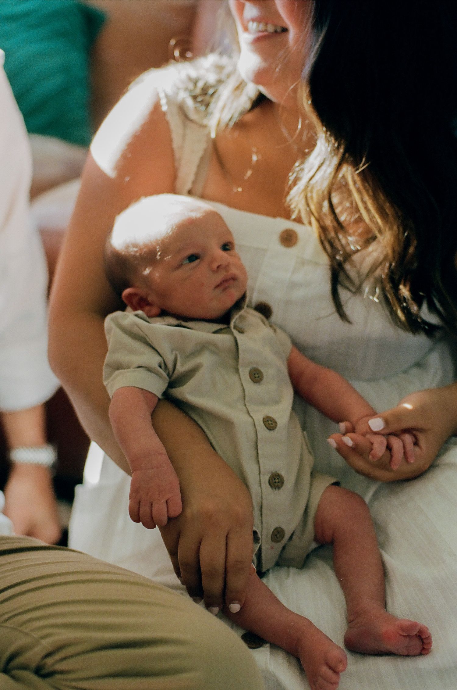 Newborn baby in sage green romper cradled in mother's arms, with a smiling woman in white dress nearby.