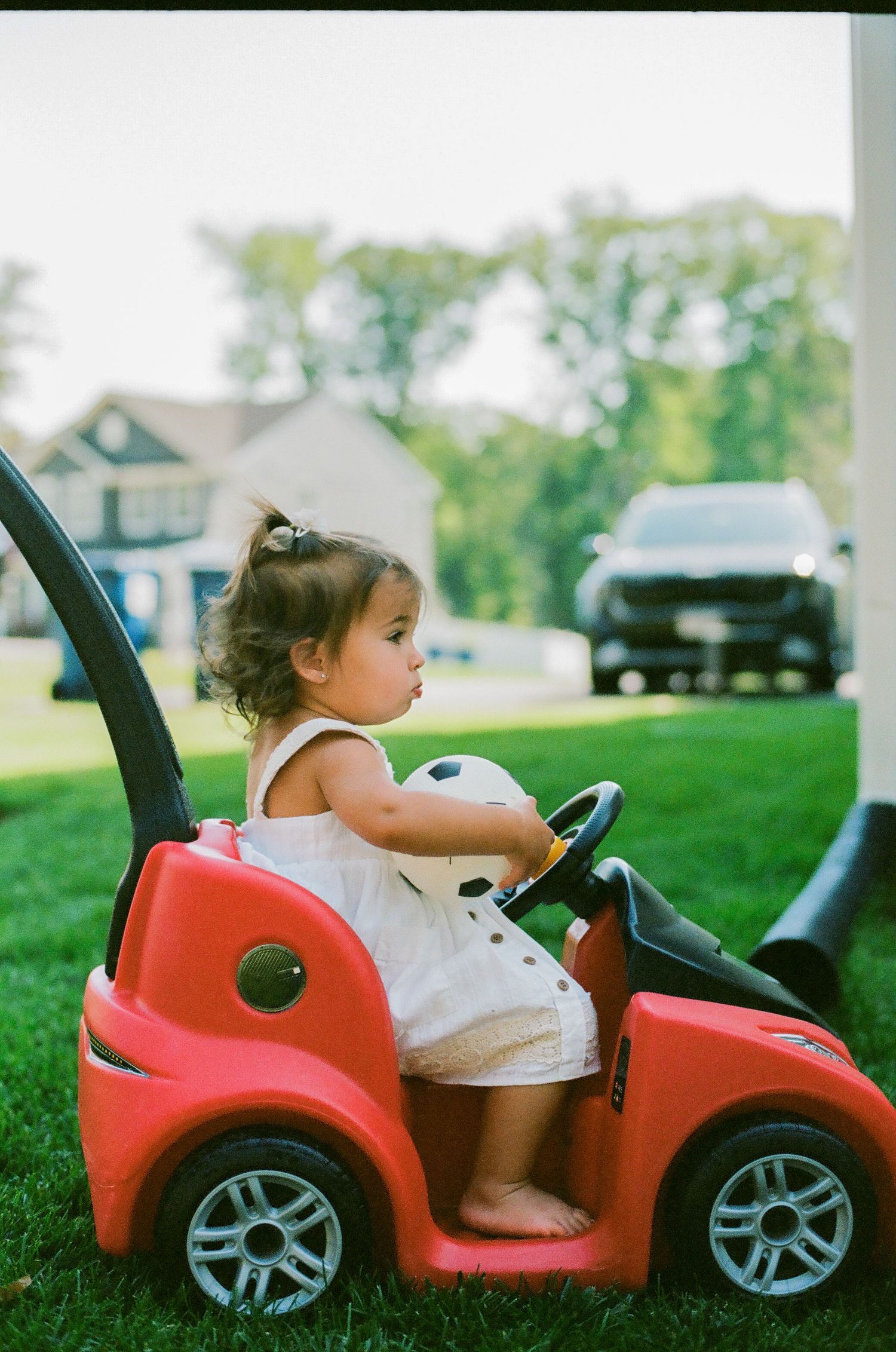 Toddler girl in white dress sitting in red toy car on green grass with suburban background.