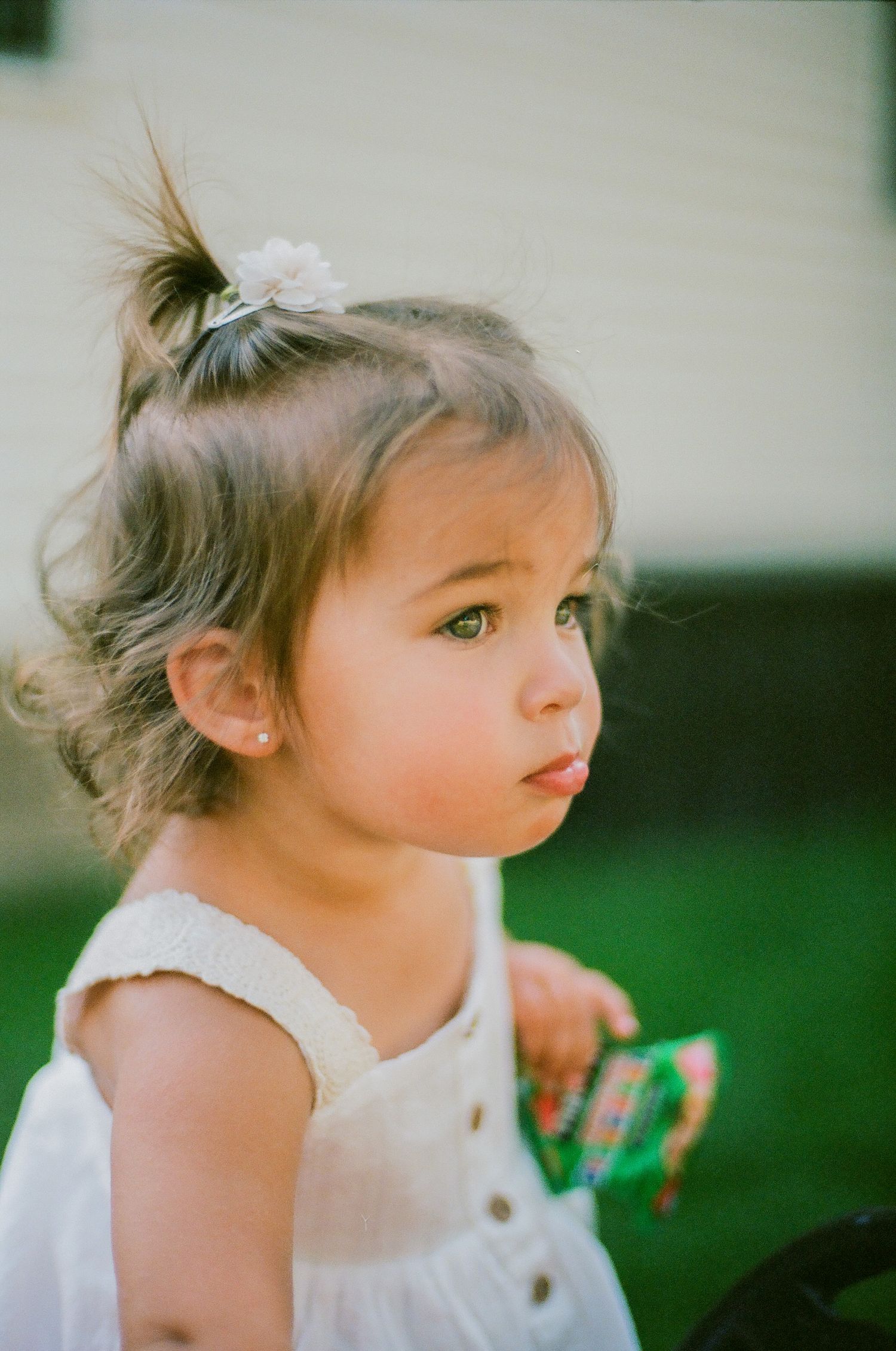 A young toddler girl in a white dress with a floral hair clip gazes curiously to the side outdoors.
