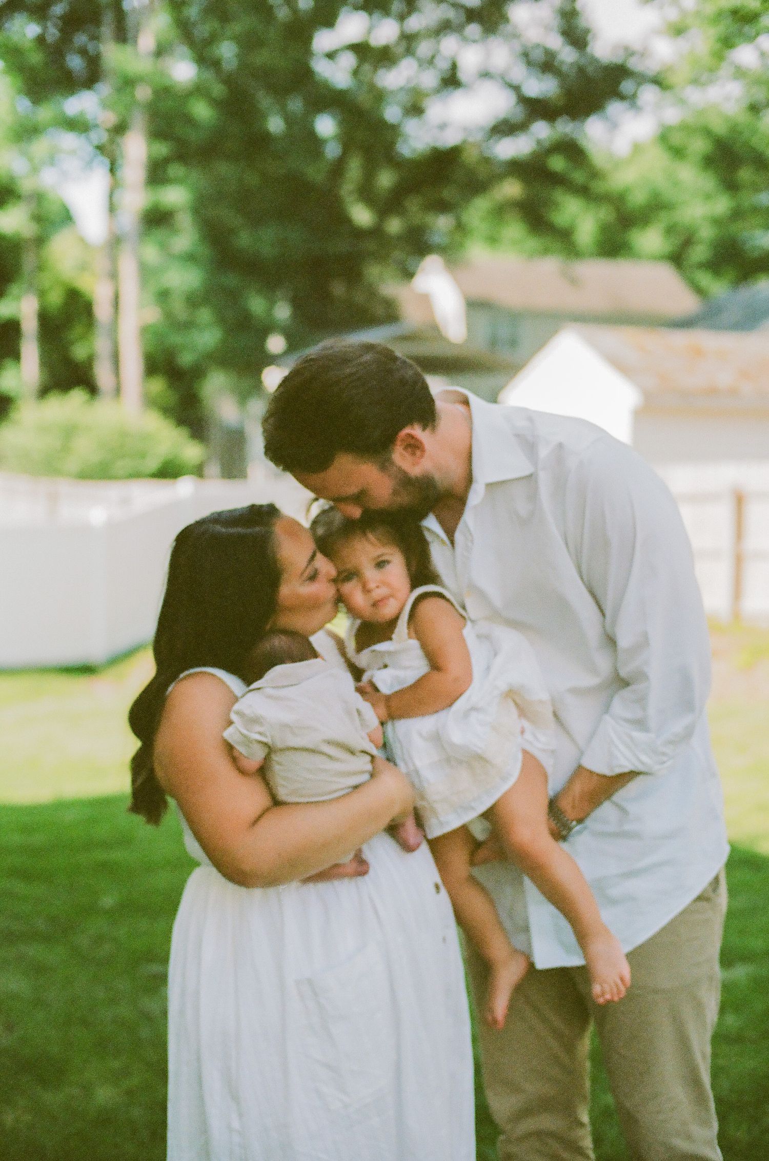 A couple in white clothing share a tender kiss on their toddler daughter's head outdoors on a green lawn.