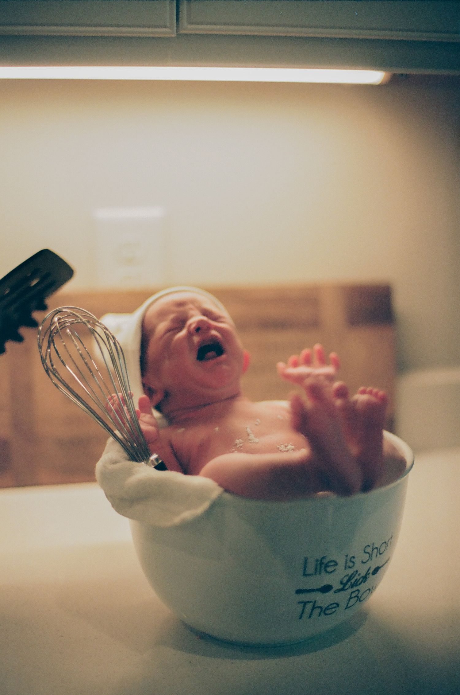 Newborn baby crying inside a large mixing bowl with a whisk, creative funny infant photo concept.