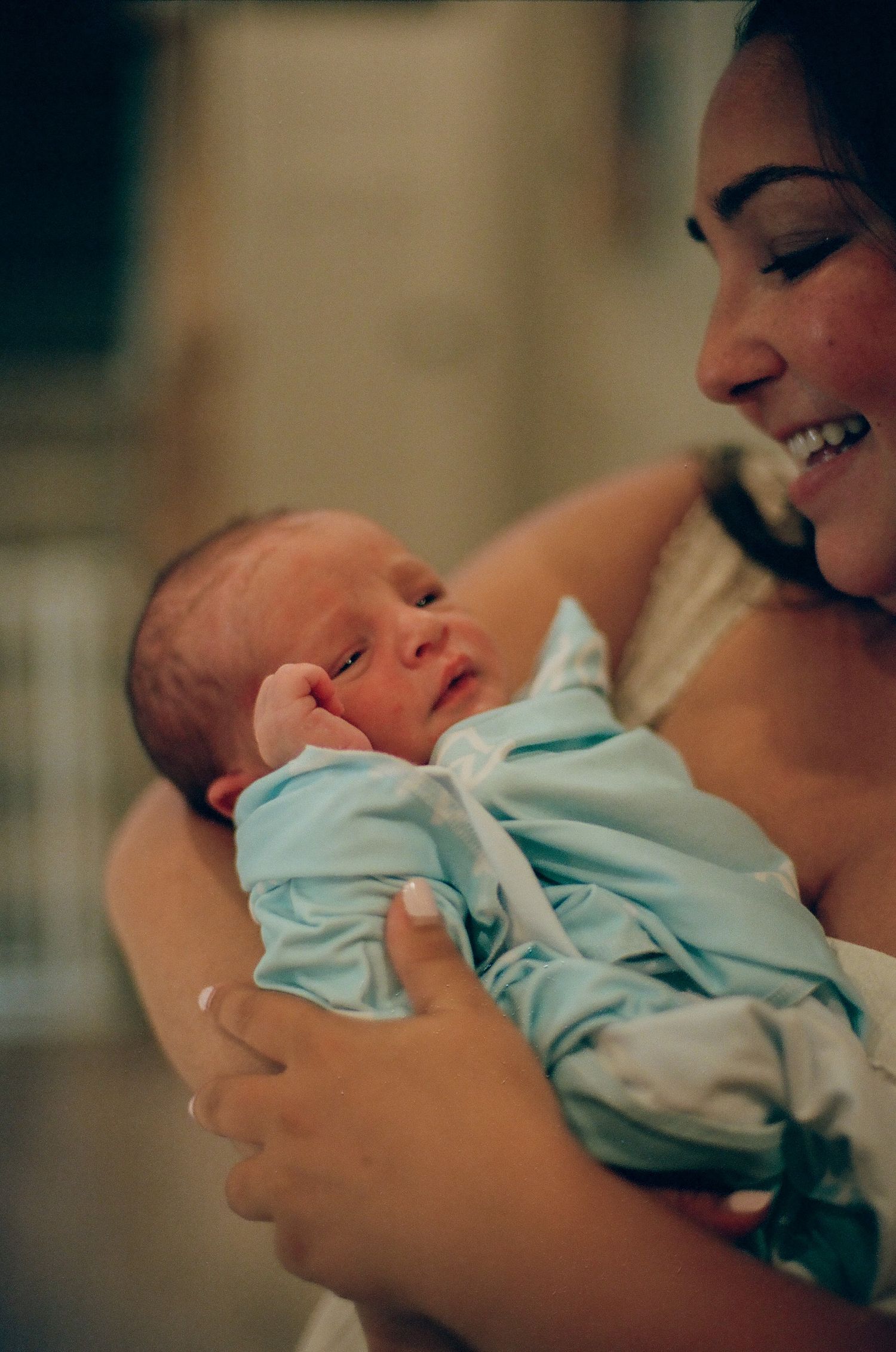 A smiling mother tenderly cradles her newborn baby dressed in a light blue outfit.