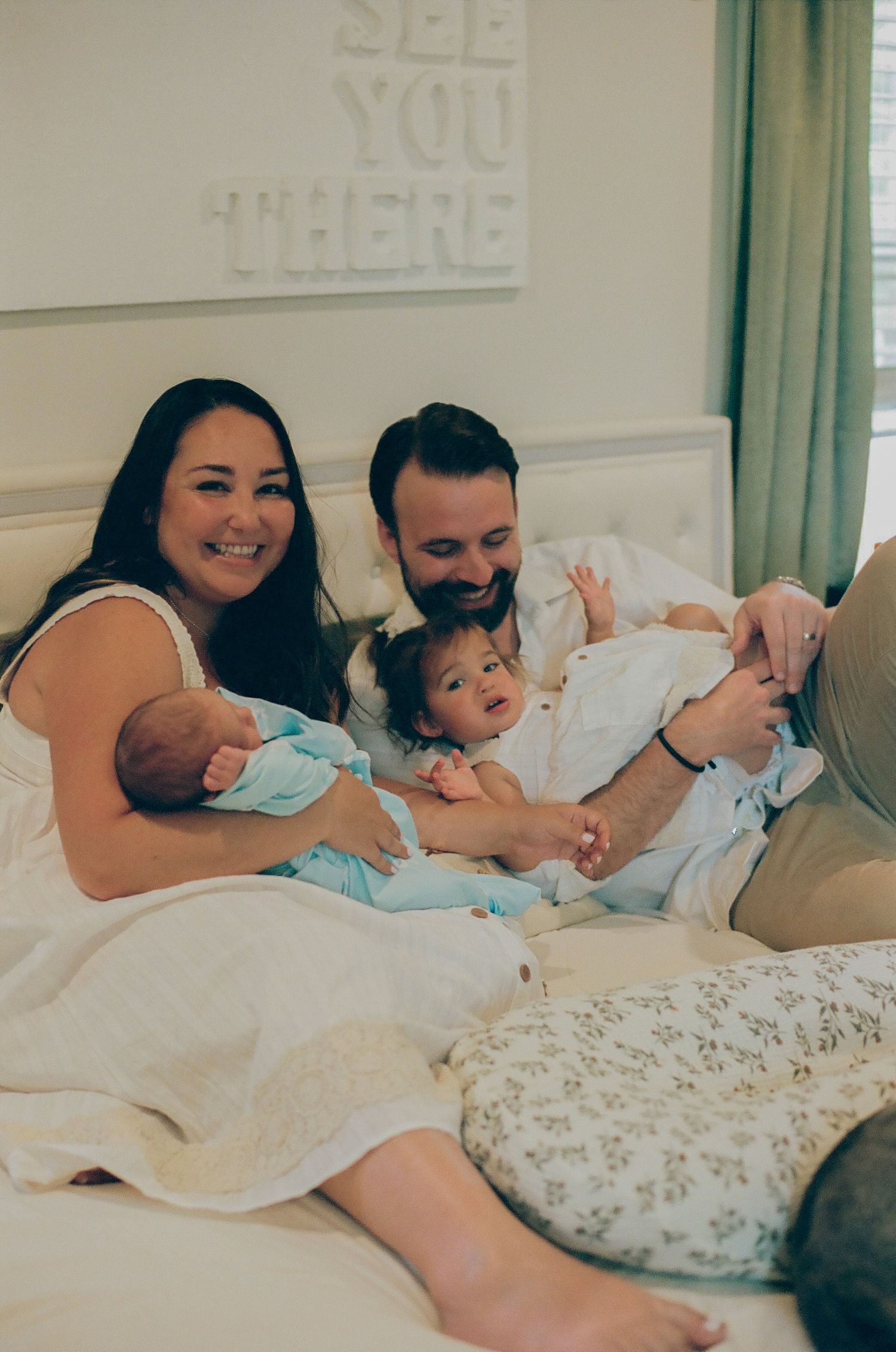 Happy family with two young children relaxing on a white bed, smiling and bonding in a bright, cozy bedroom.