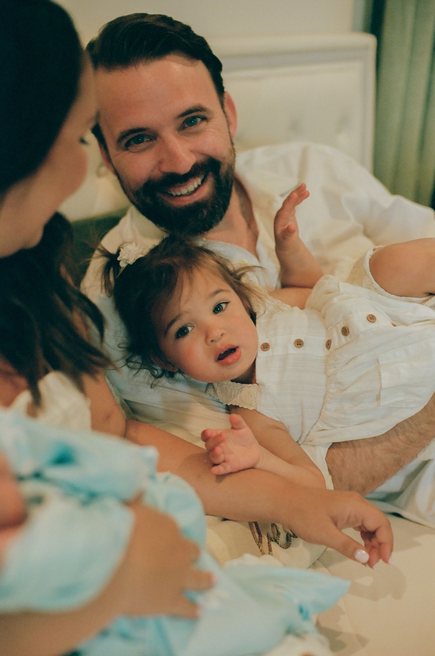 Smiling bearded man holding a toddler on a bed while a woman cradles a newborn baby nearby.