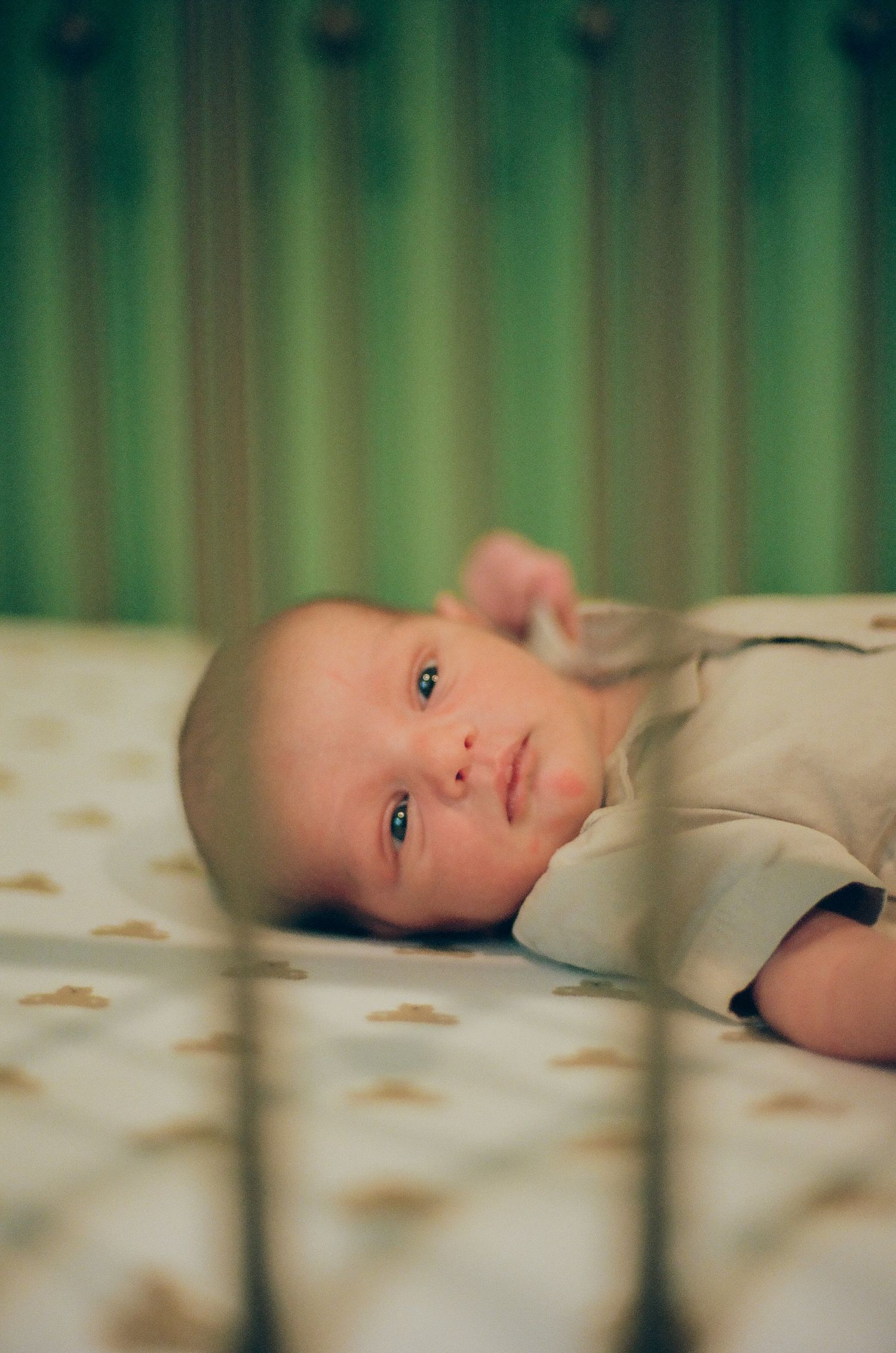 A newborn baby with blue eyes lying awake in a crib on a dotted mattress with green striped wallpaper background.