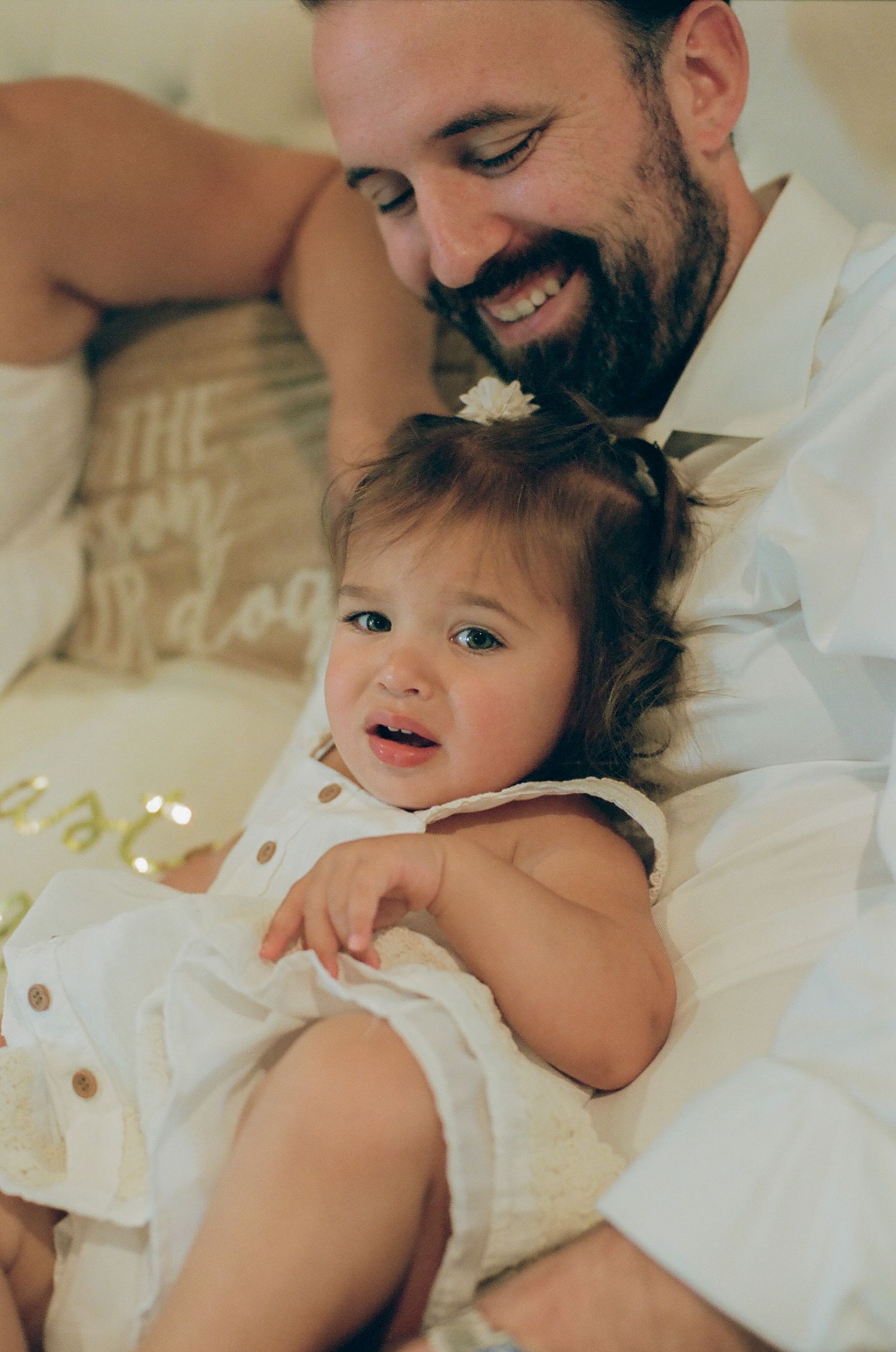 Smiling bearded father cuddling happy baby girl in white dress on a cozy bed.
