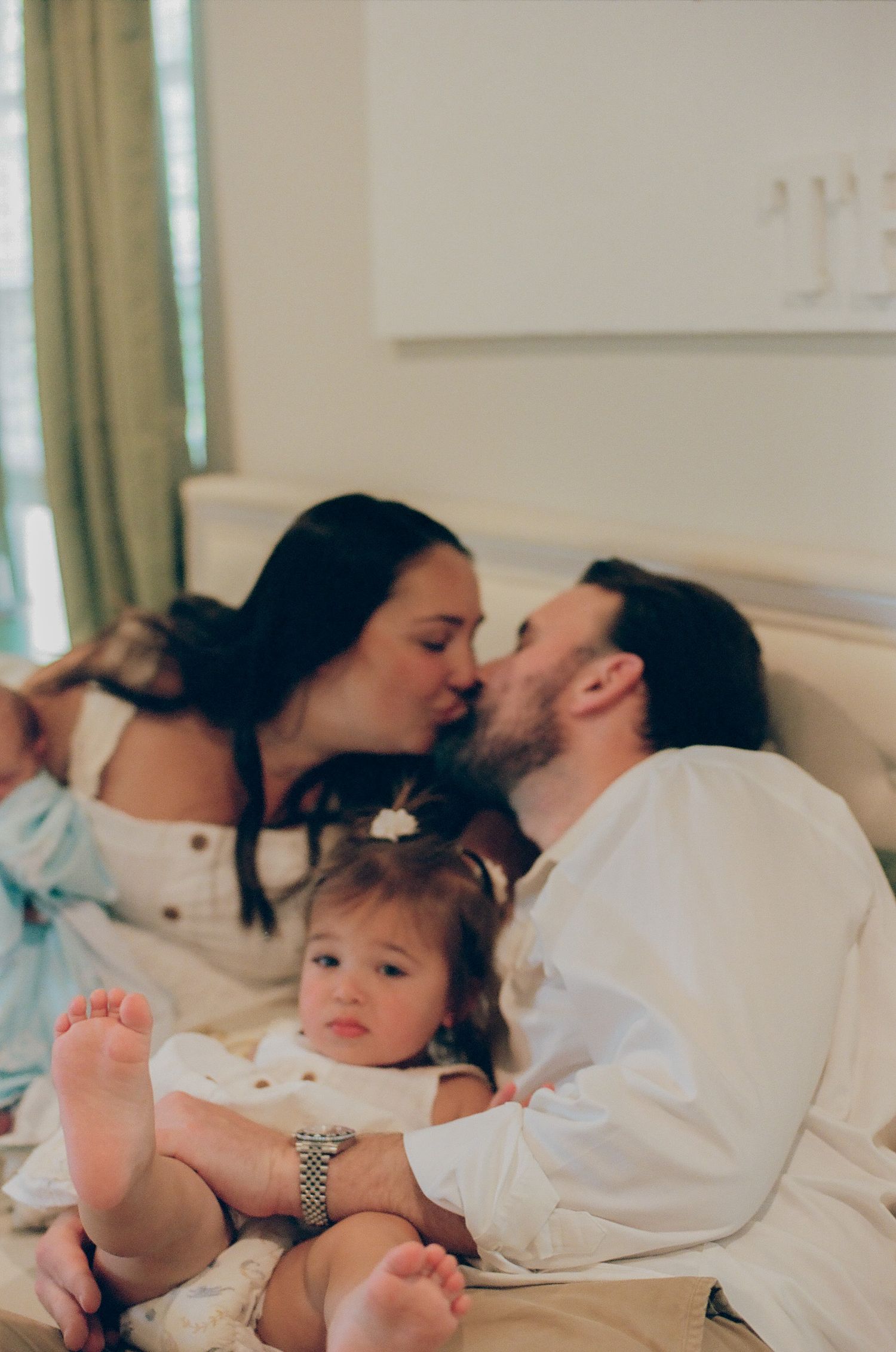 A couple kisses on a bed while their baby girl sits between them in a cozy bedroom setting.