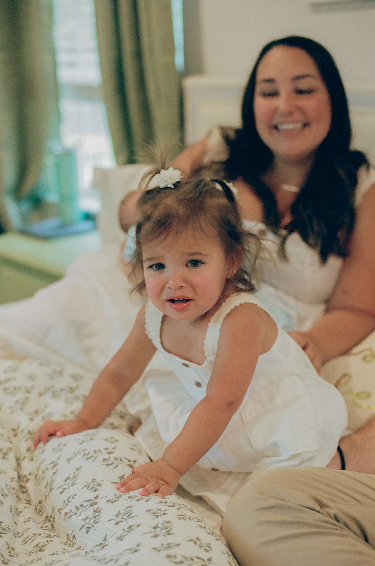 A smiling toddler girl in a white dress sits on a bed while a woman in white smiles warmly behind her.