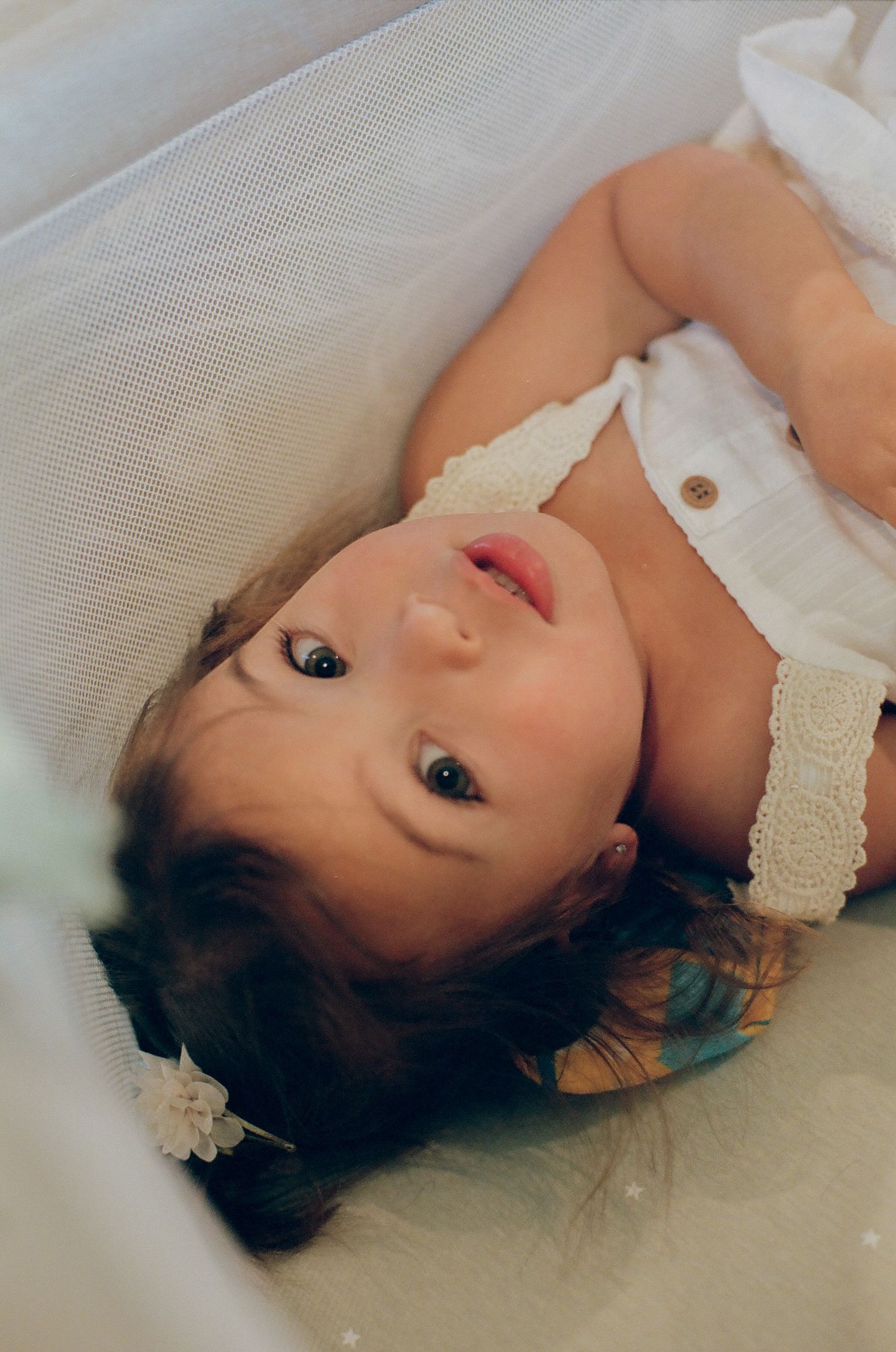 A baby girl with dark curly hair and a white lace outfit lying down, looking up with bright curious eyes.