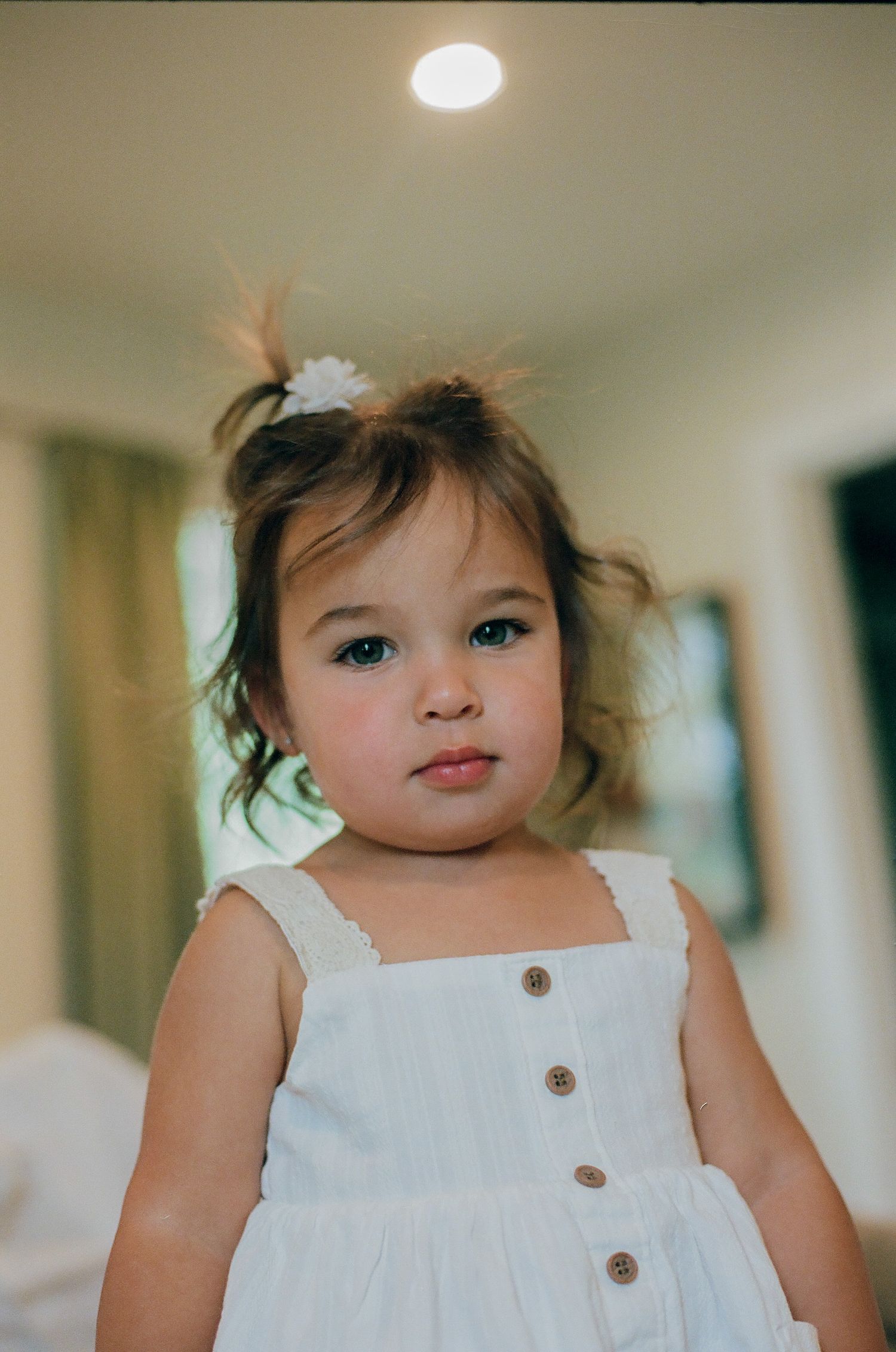 Adorable toddler girl in white button dress with flower hair clip poses indoors with a serious expression.