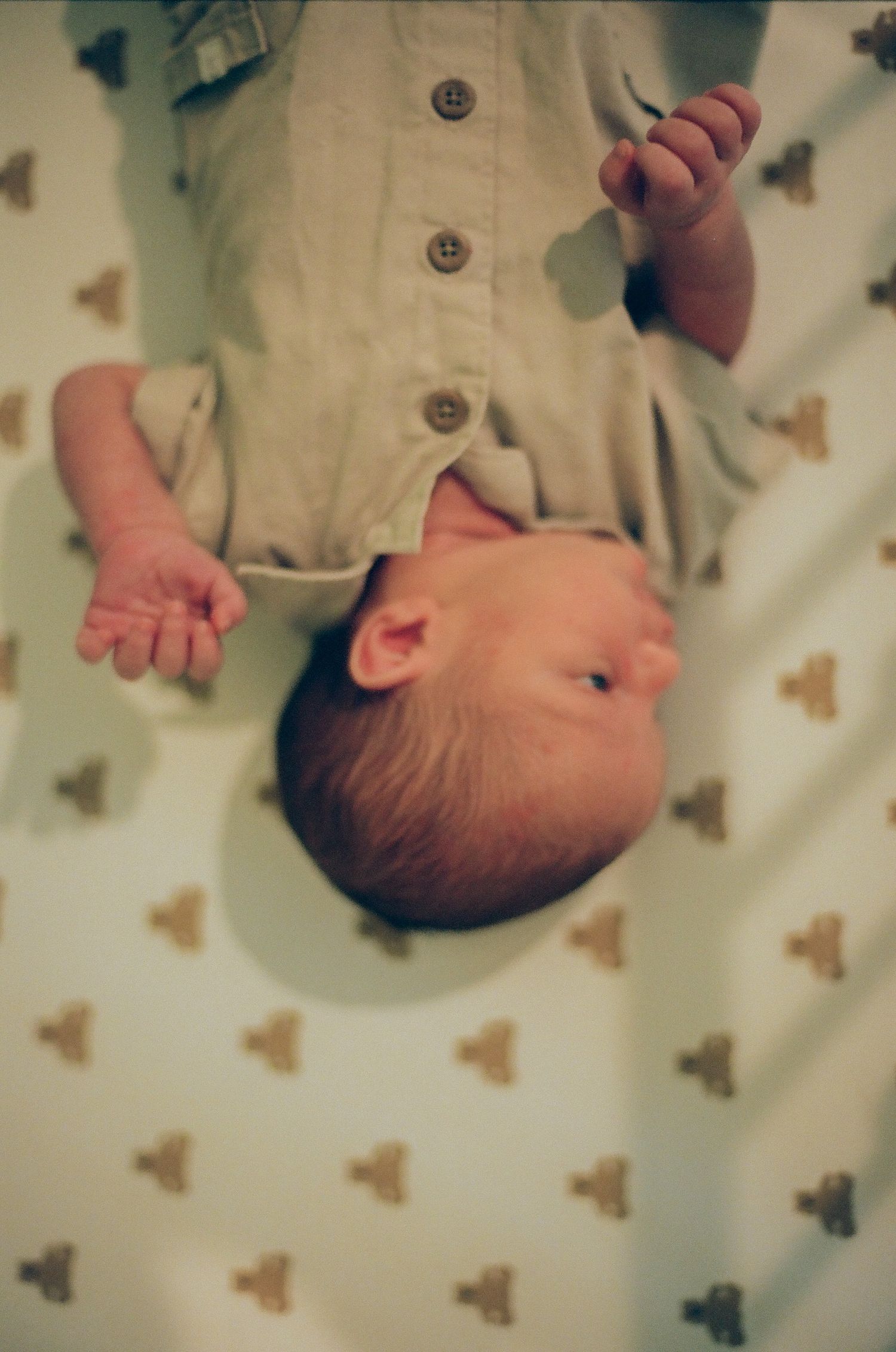 Newborn baby lying on back in crib wearing olive green button-up onesie on patterned white sheet.