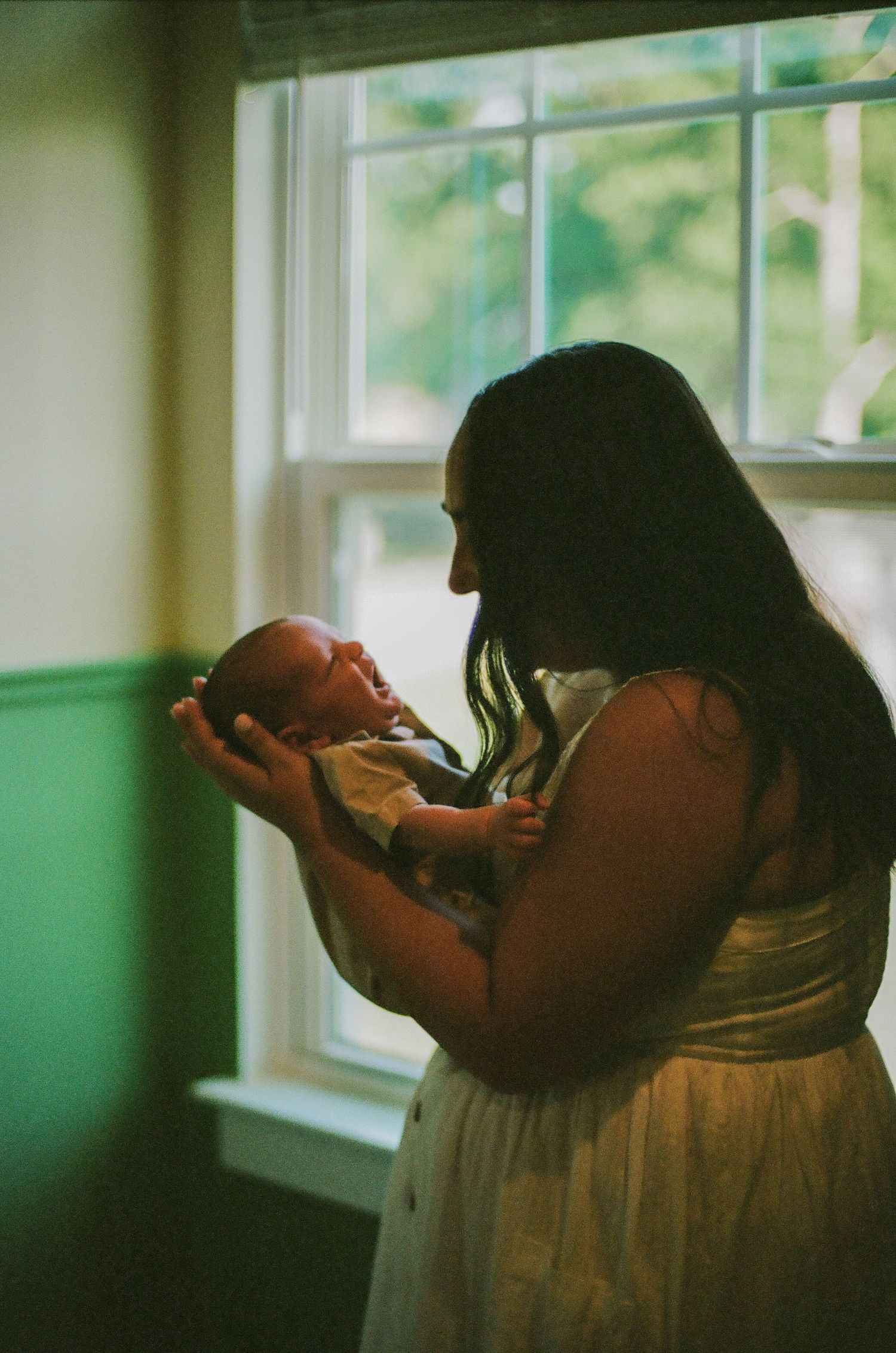 A mother tenderly holds her newborn baby near a sunlit window in a heartfelt film photography portrait.