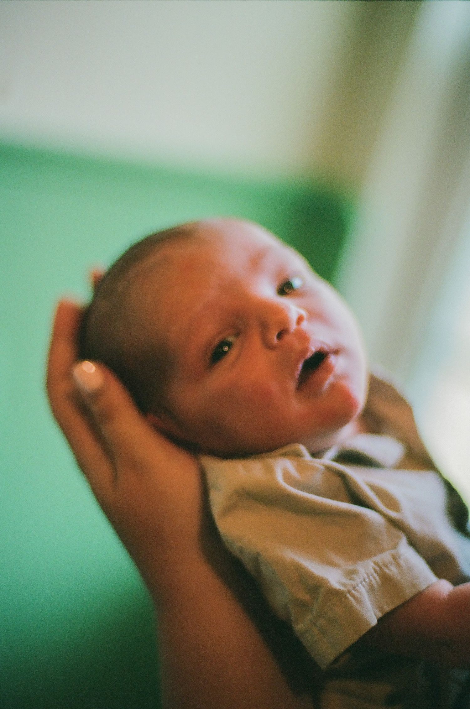 A newborn baby with wide eyes is gently cradled in an adult's hands against a soft green background.