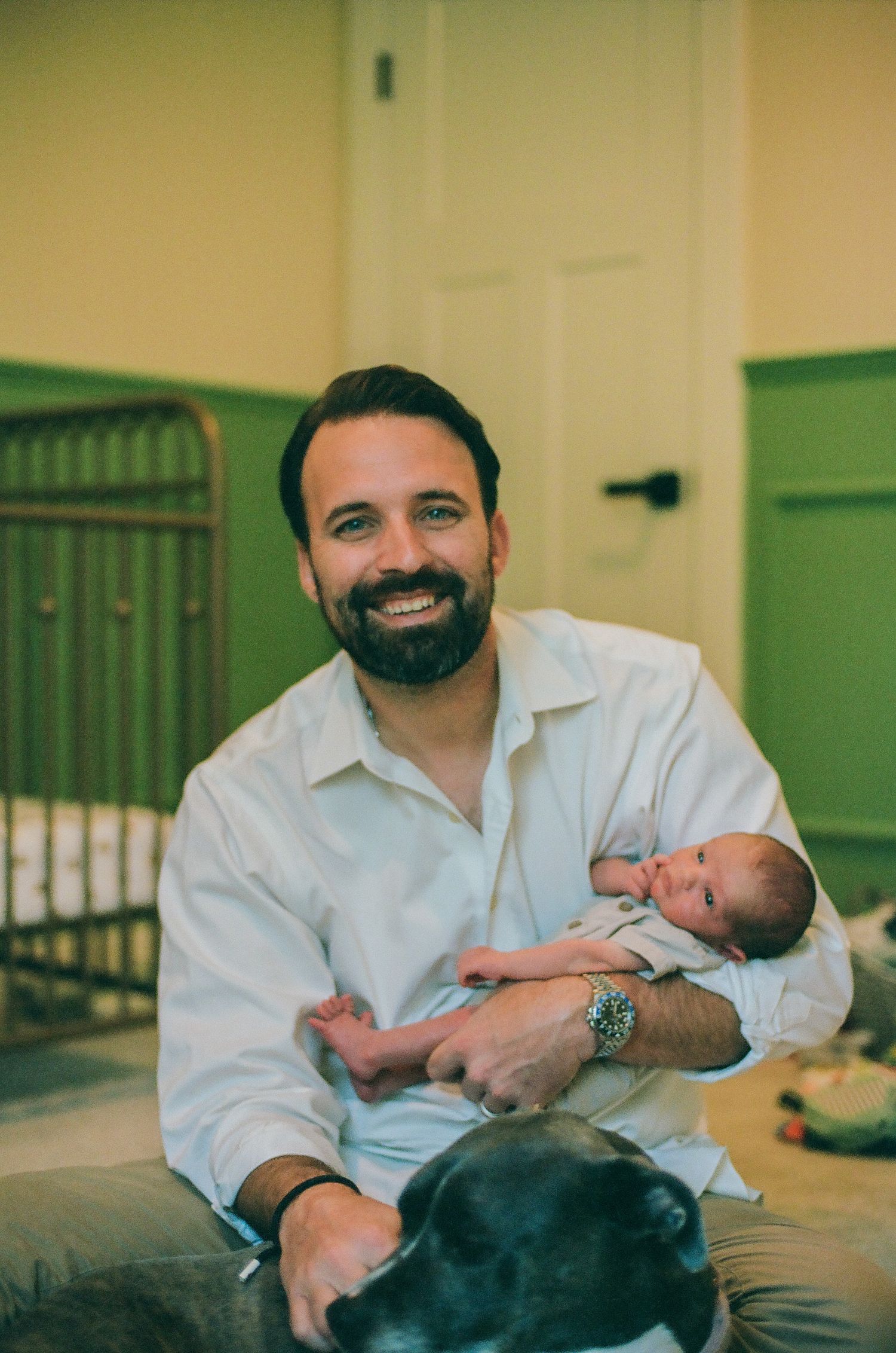 Smiling bearded man holding a newborn baby with a black dog in foreground, sitting in a nursery room.