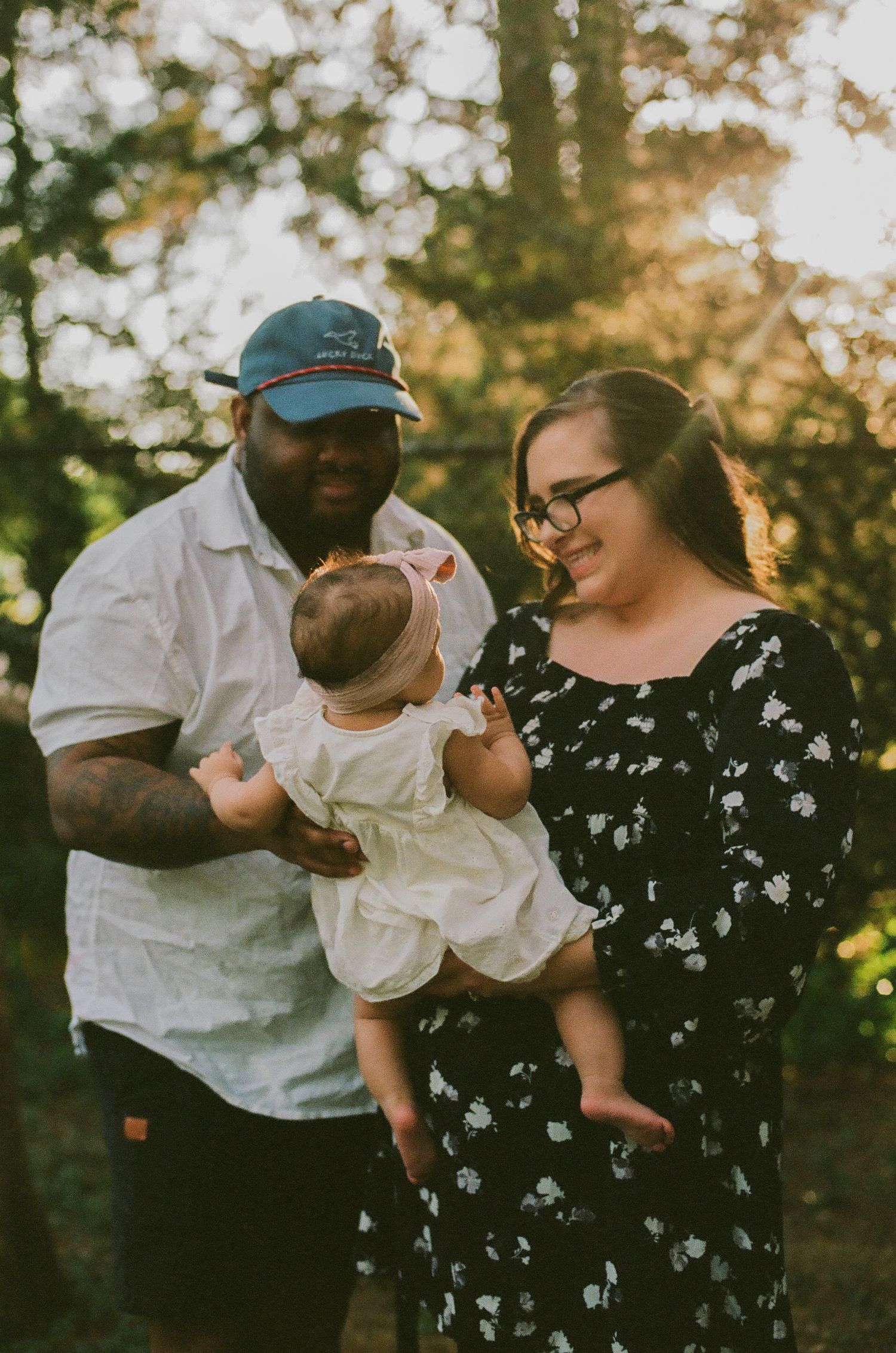 A happy family poses outdoors at golden hour, father holding baby in white dress beside smiling mother in floral dress.