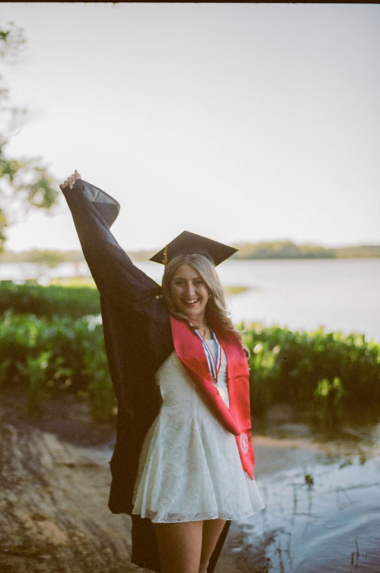 Smiling graduate in cap and gown with red sash poses outdoors by a lake holding her cap up in celebration.