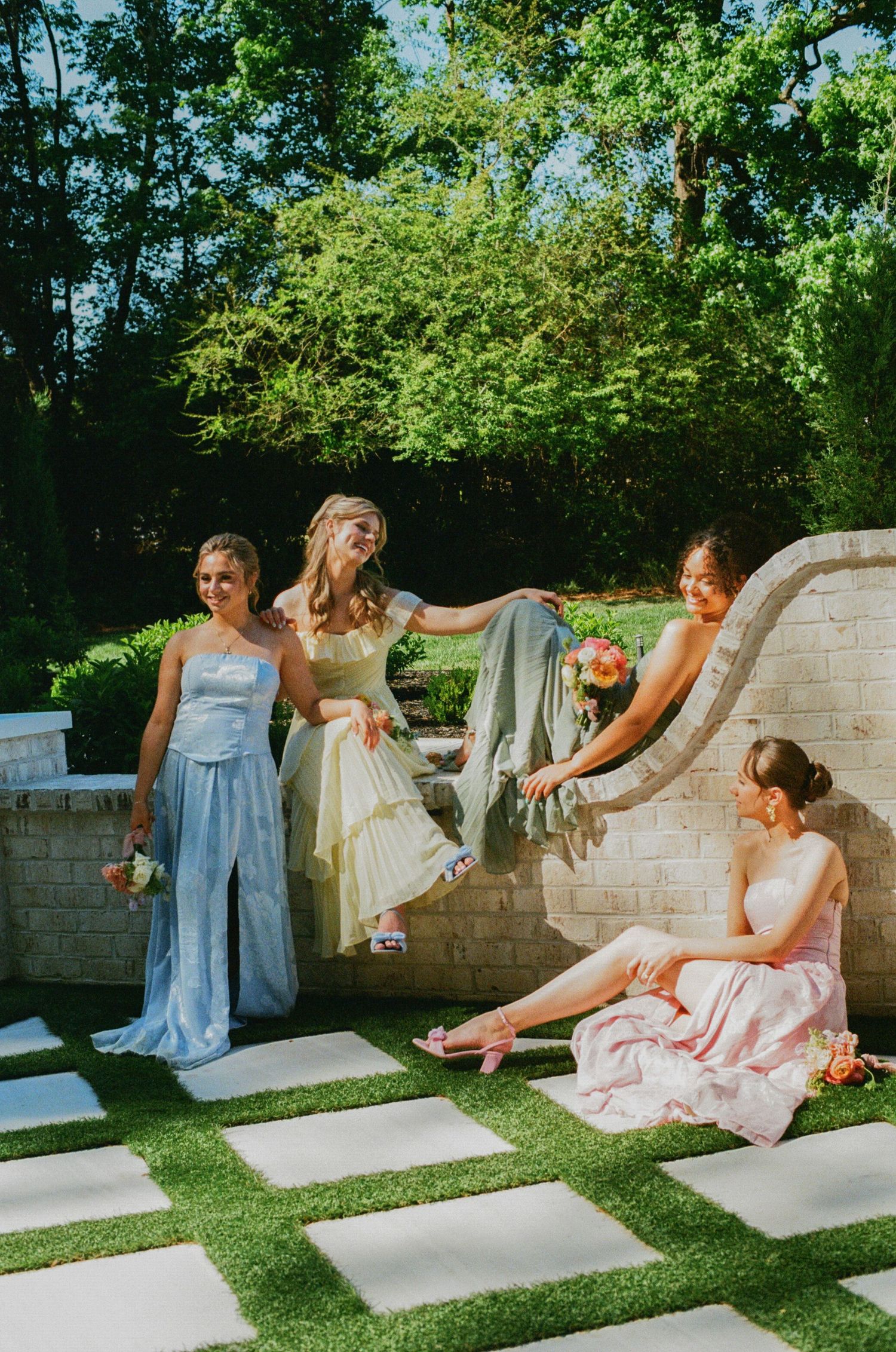Four young women in elegant pastel formal dresses pose outdoors in a garden setting on a sunny day.