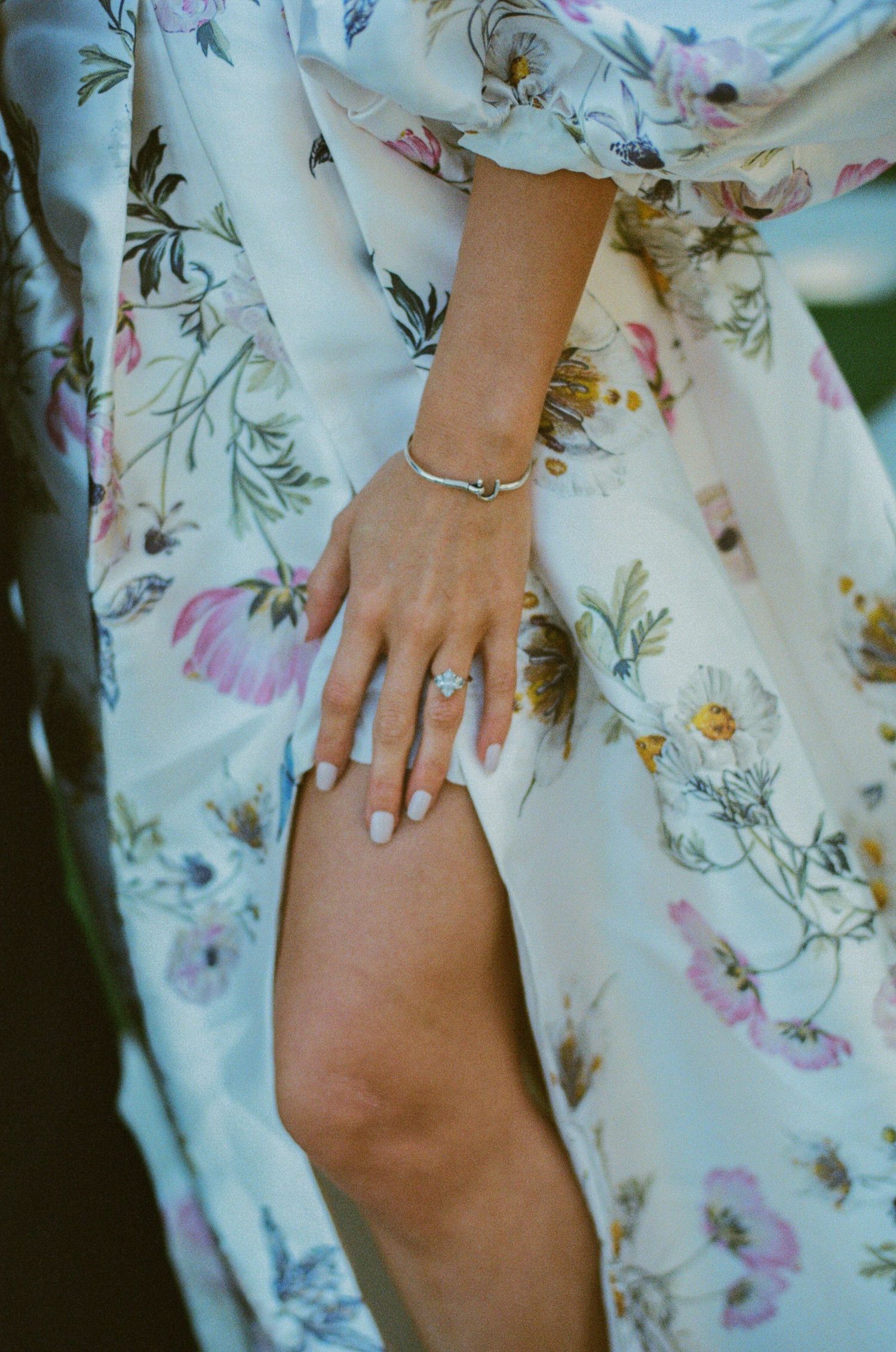 Close-up of a bride's hand with engagement ring and bracelet resting on floral wedding dress.