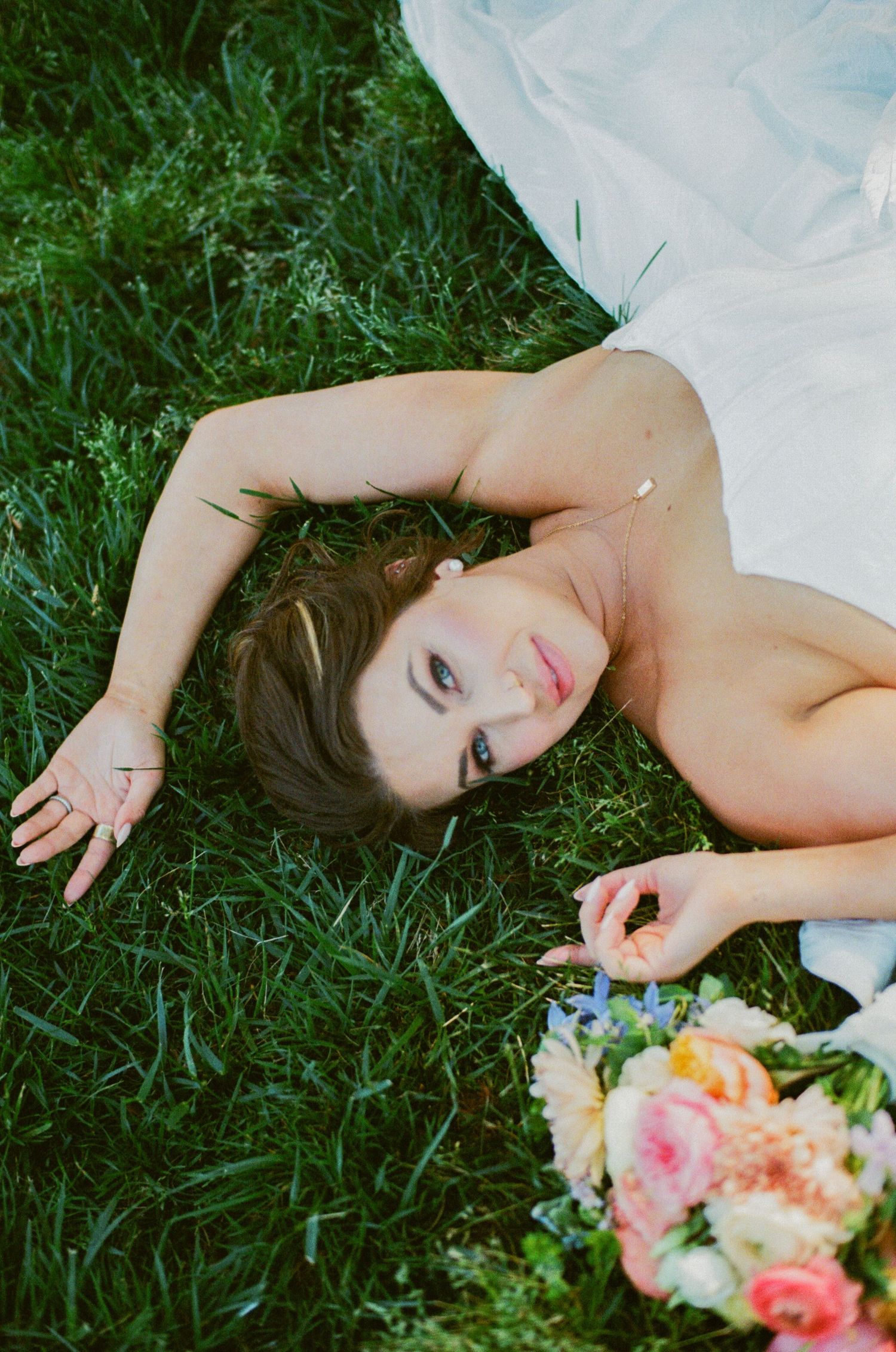 Bride in white dress lying on green grass holding a colorful bouquet of pink and peach flowers, looking up at camera.