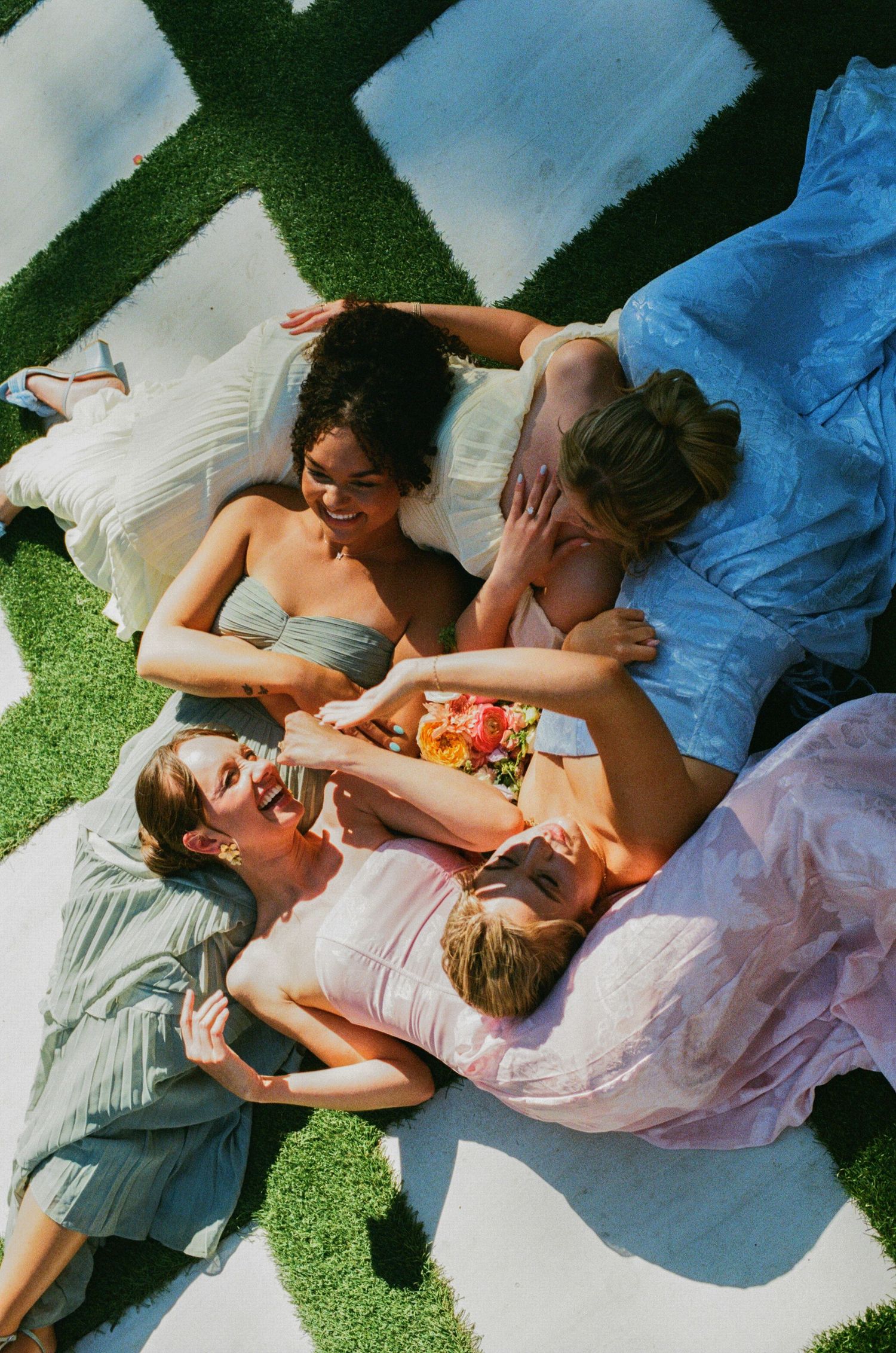 Four women in pastel dresses laughing and lying together on a white blanket on green grass.