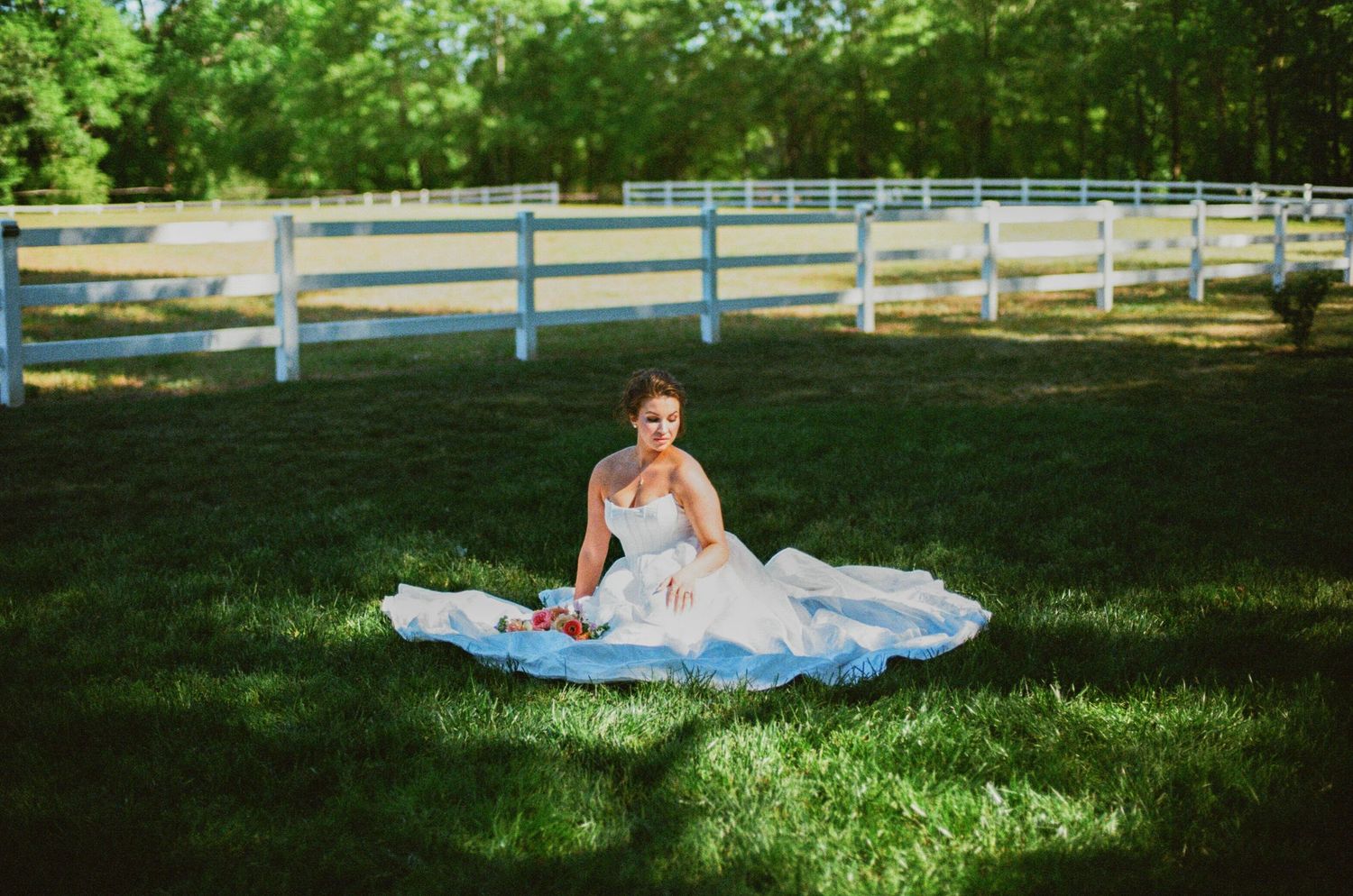 Bride in white wedding gown sitting gracefully on green grass near white fence at outdoor farm venue.