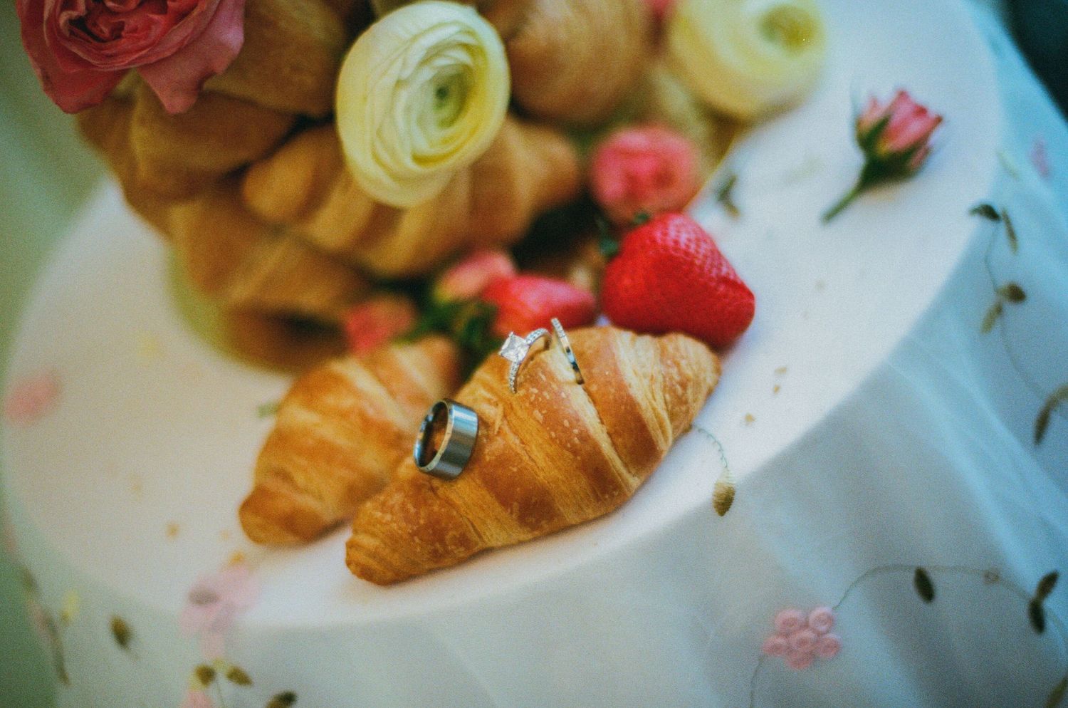 Wedding rings resting on golden croissants beside fresh strawberries and roses on a white tiered cake.