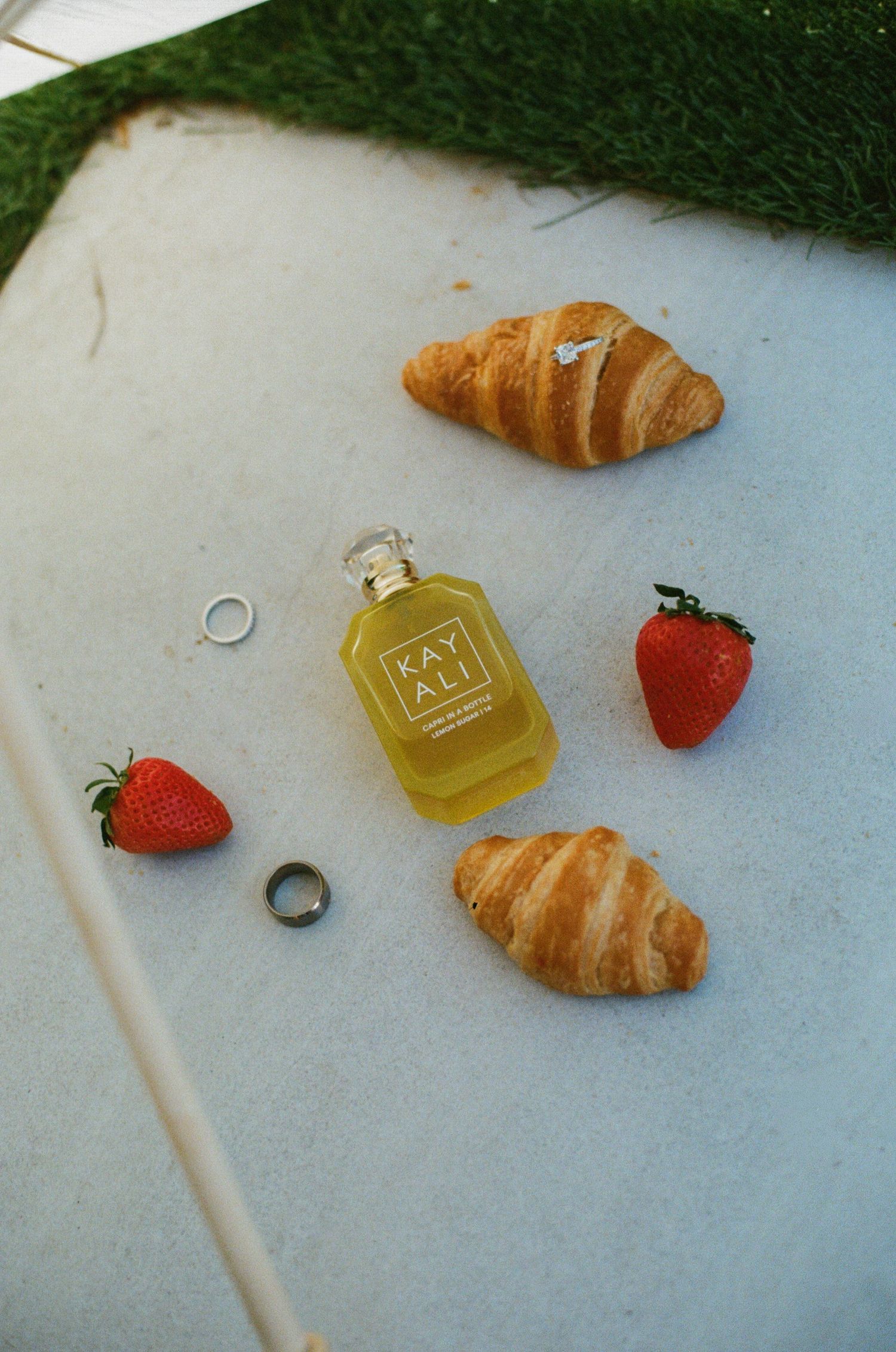 Kayali perfume bottle surrounded by croissants, strawberries, and rings on a white surface with green grass border.