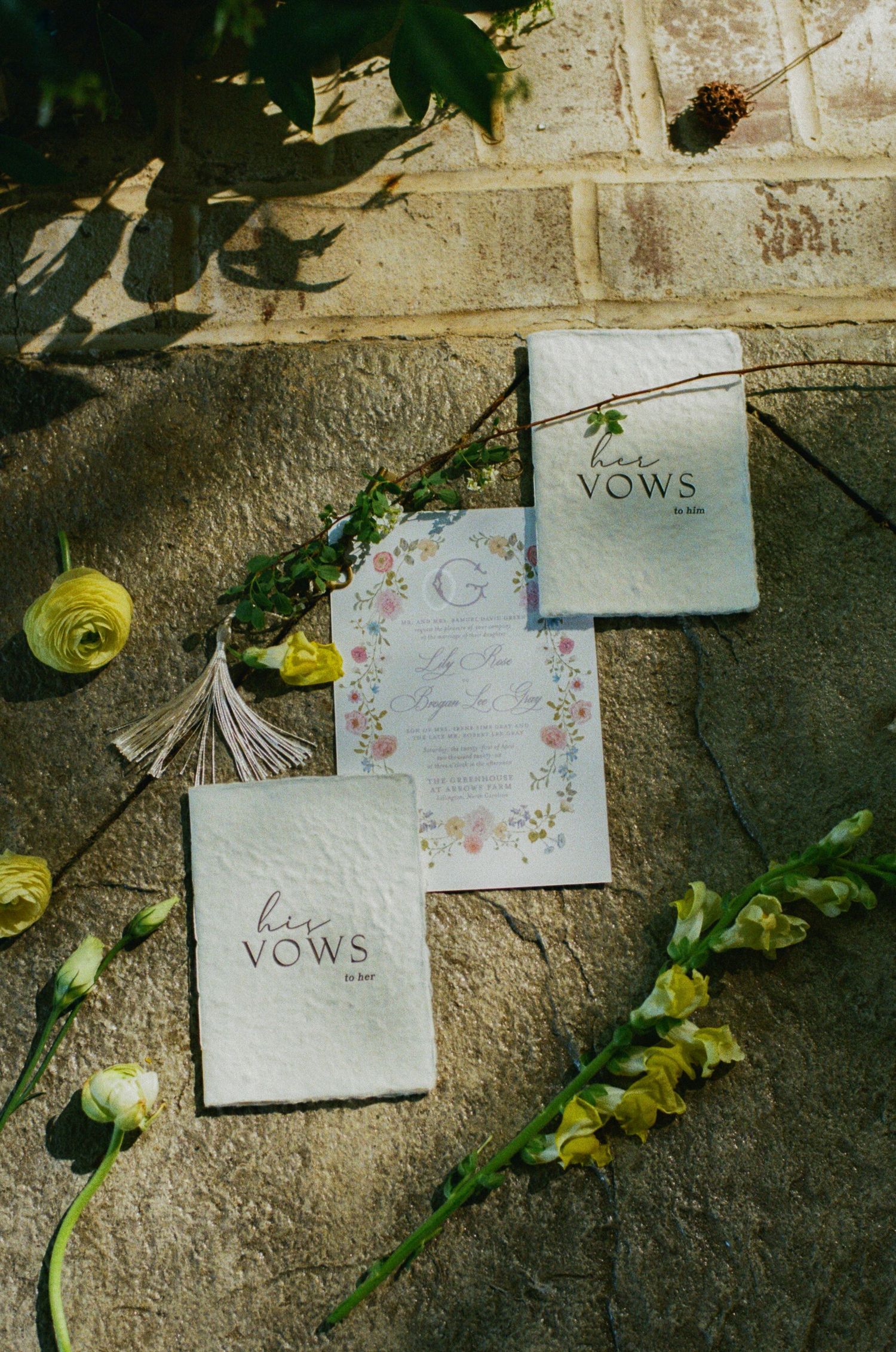 Elegant wedding vow booklets and floral invitation suite arranged on stone surface with yellow flowers and greenery.