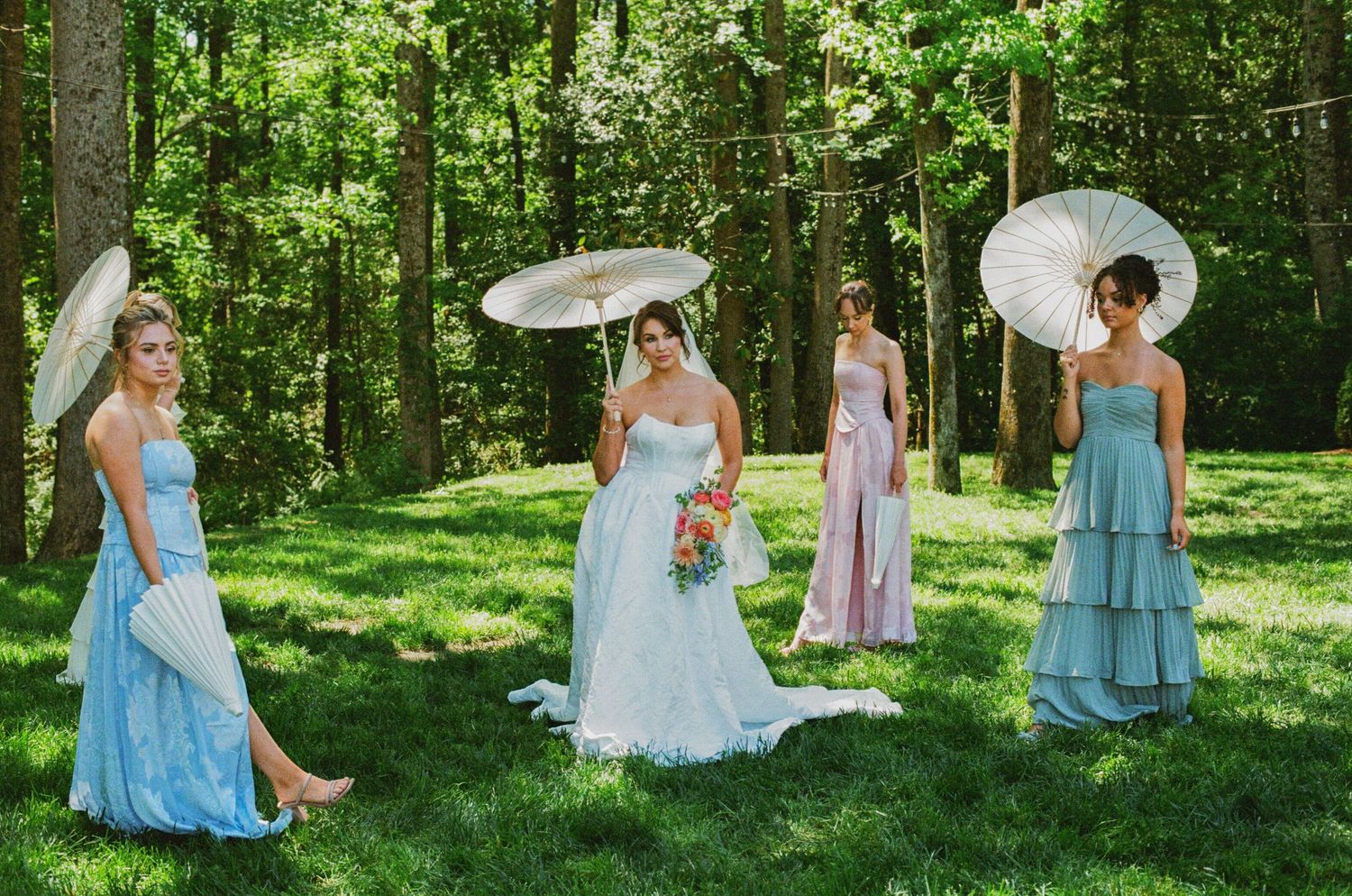 Bride in white gown with bridesmaids in blue and pink dresses holding parasols outdoors in lush green forest setting.