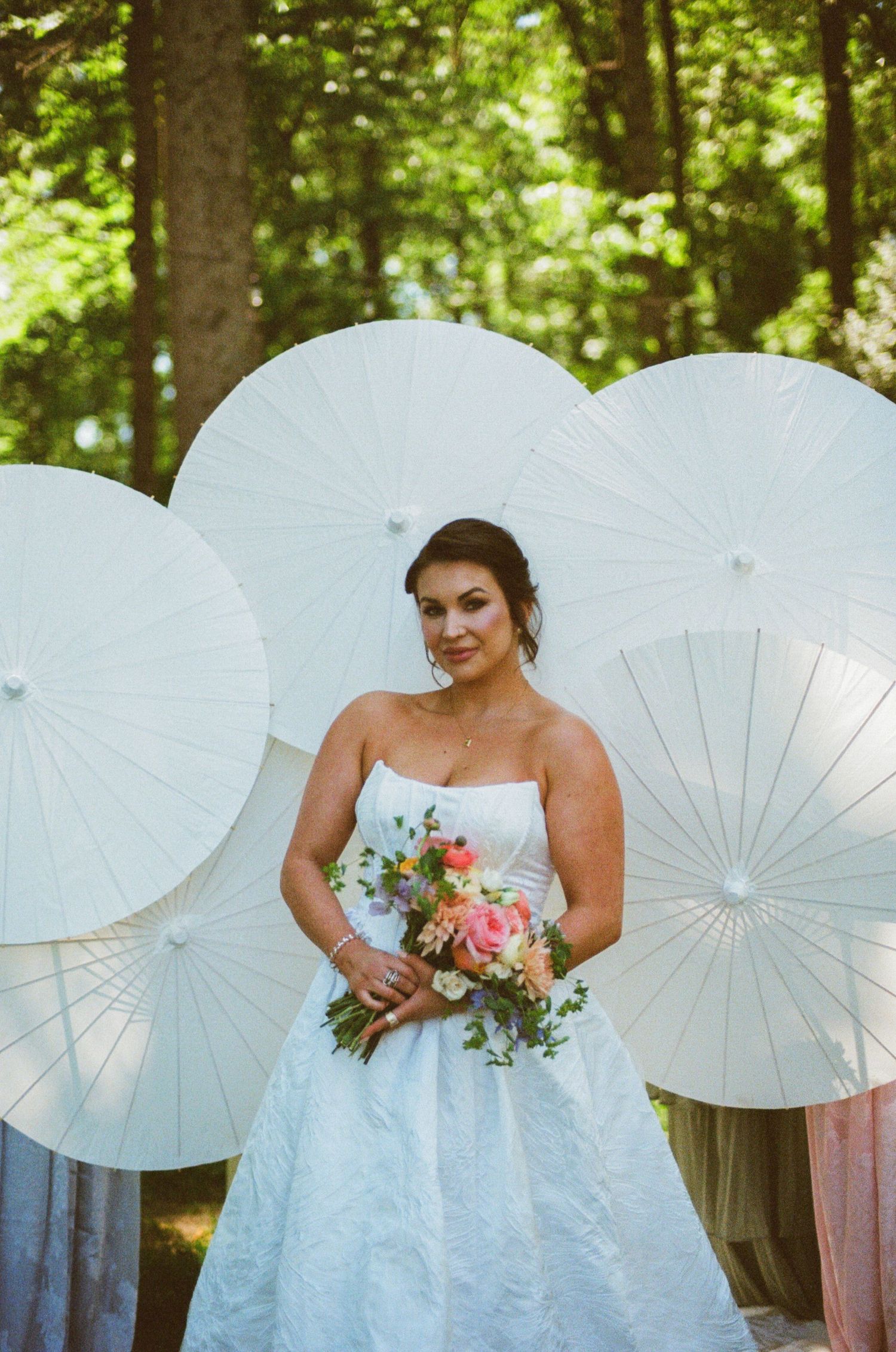 Bride in strapless white gown holding colorful bouquet, posing before white paper parasols in a lush forest setting.