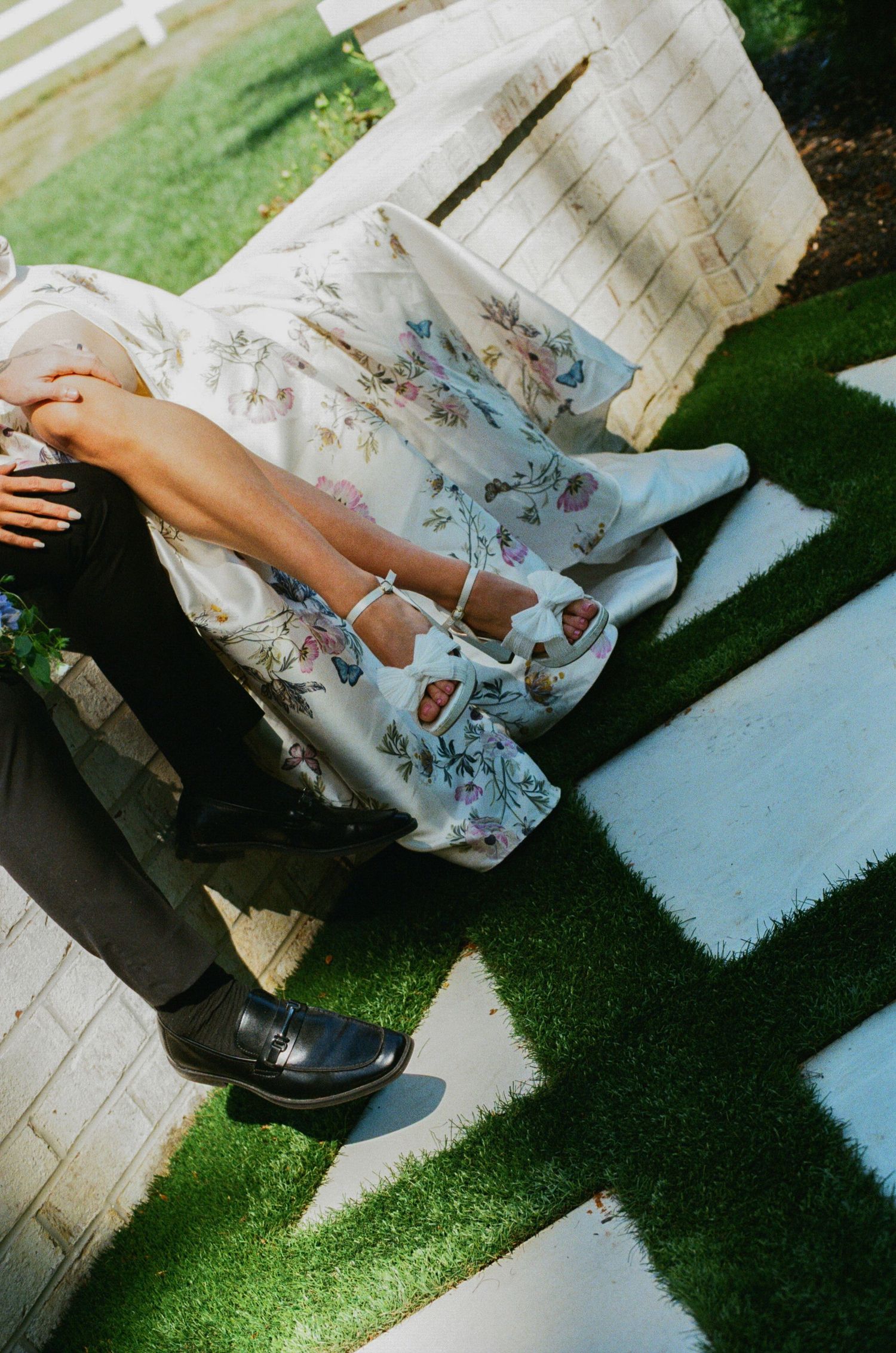 Couple sitting outdoors, woman in floral dress with blue strappy heels beside man in dark pants and black dress shoes.