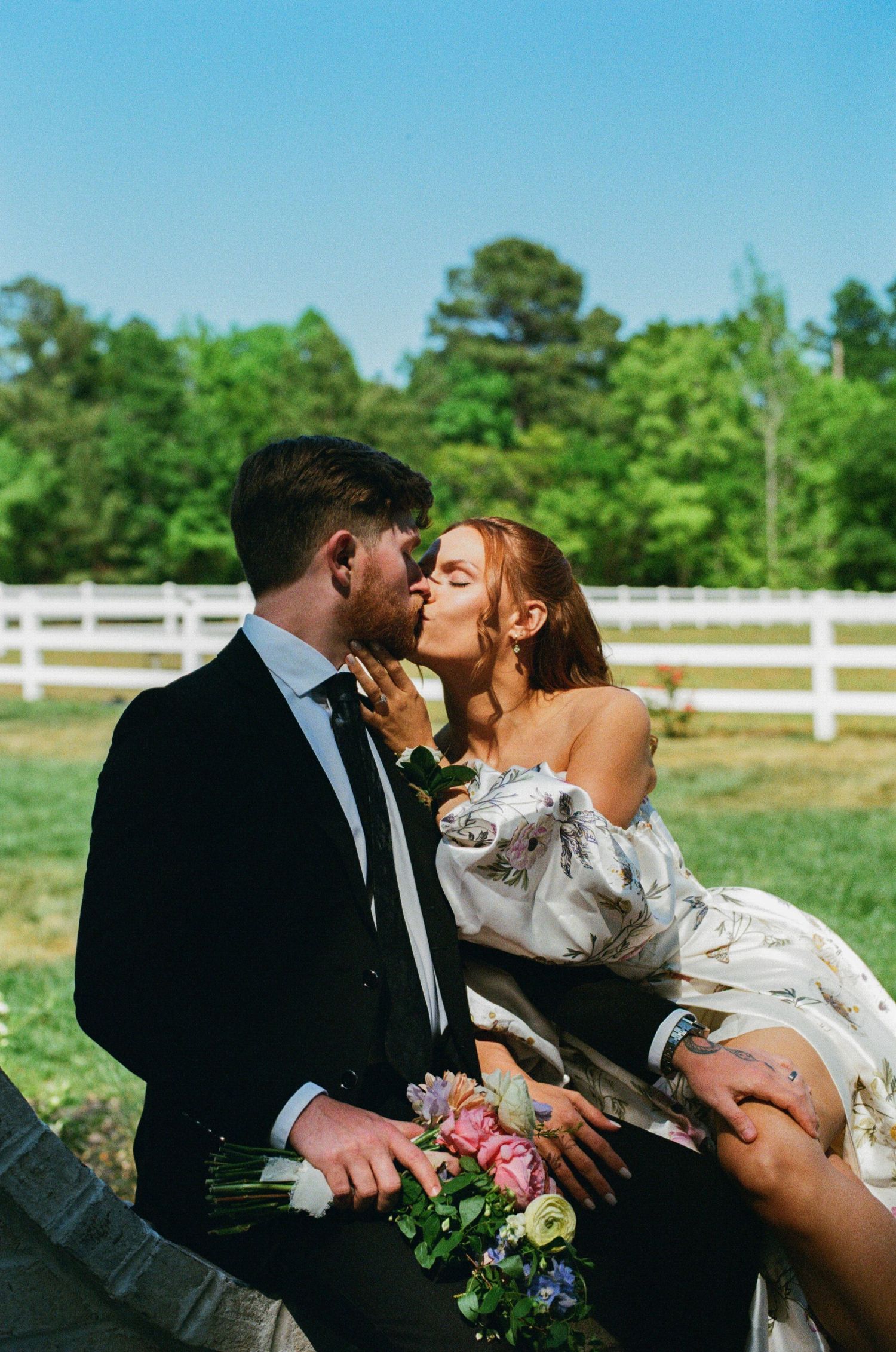 Bride and groom sharing a romantic kiss outdoors at a farm wedding venue, with white fence and green trees.
