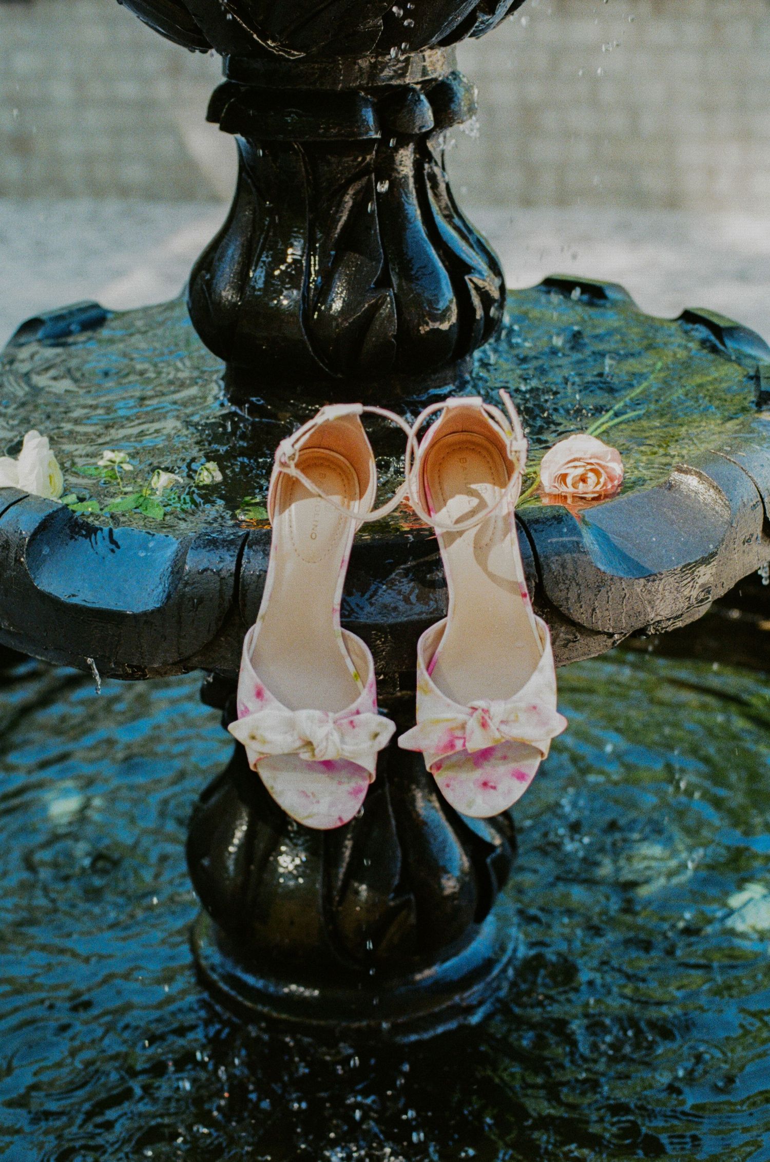 Floral bow heeled sandals displayed on a vintage black water fountain with a pink rose, perfect bridal wedding shoes.