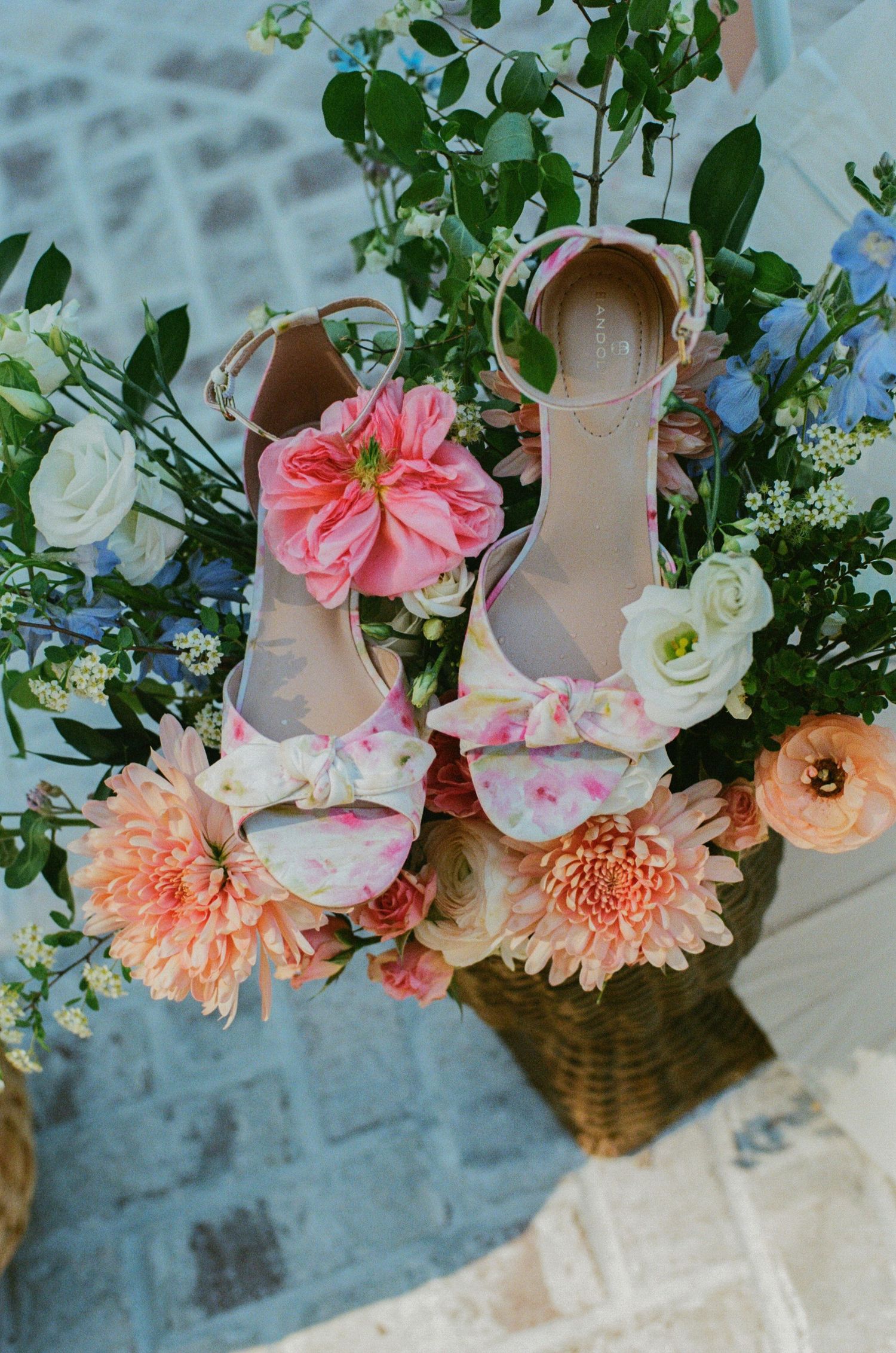 Floral heeled sandals with bow detail resting in a basket of pink, white, and blue flowers for a wedding.