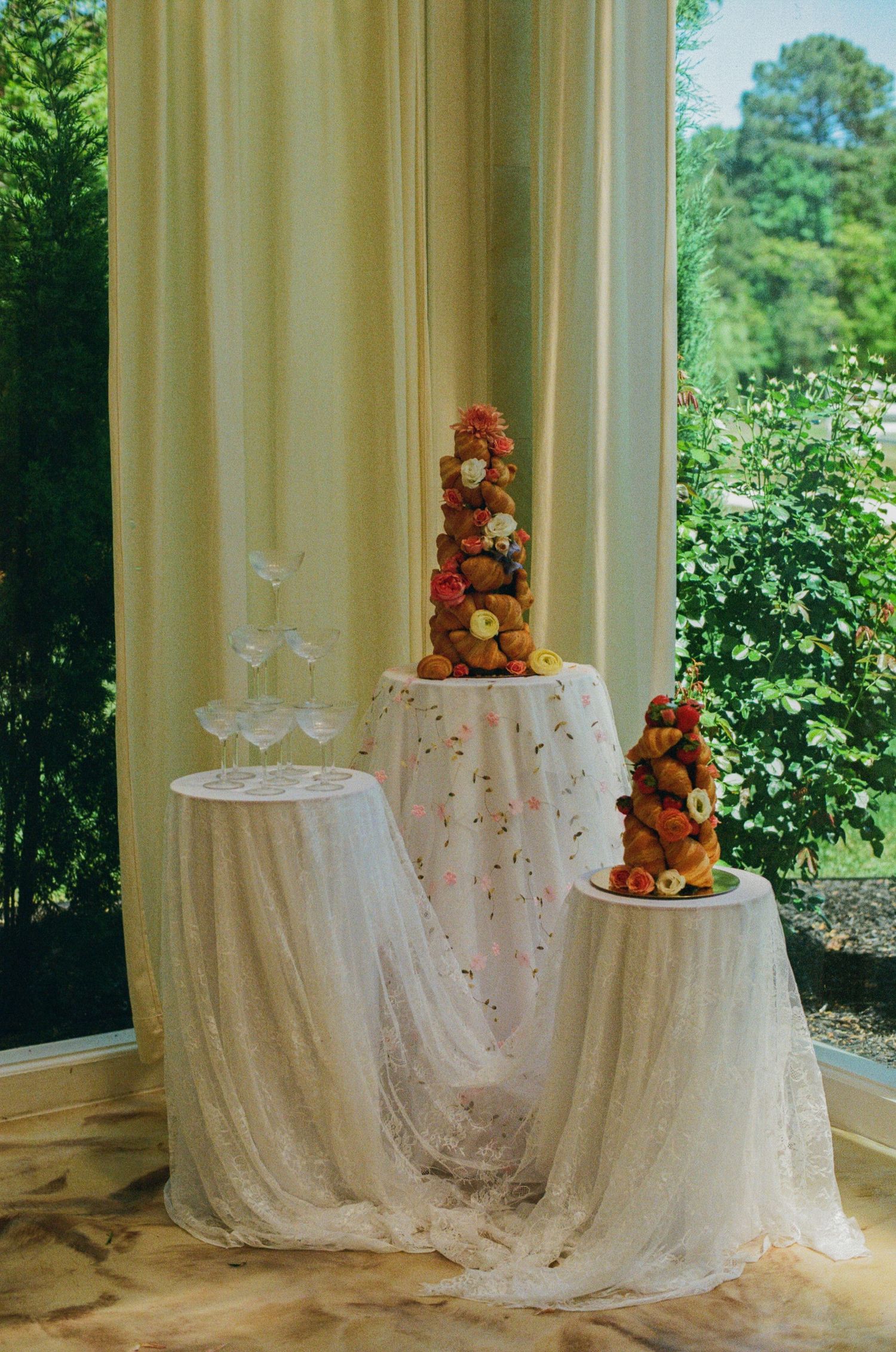 Elegant croquembouche wedding cake towers adorned with fruit and flowers on white lace draped tables near window.
